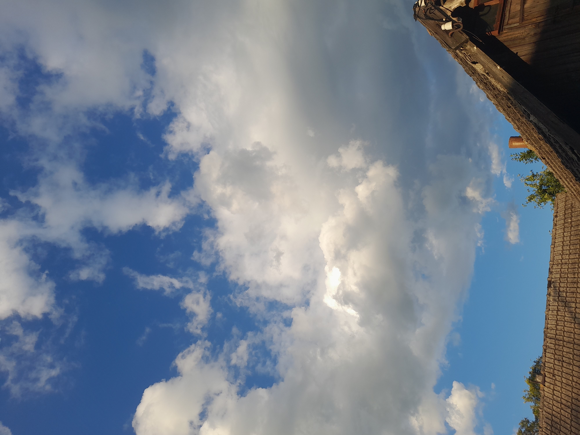 The image captures a sky filled with white and gray clouds against a blue sky. In the foreground, the tiled roof of a weathered wooden building is visible, with a chimney and some electrical fixtures on the roof. Trees can be seen peeking above the roofline. The scene appears to be in Kliushnykivka, Ukraine, with the natural light suggesting it was taken during the daytime.