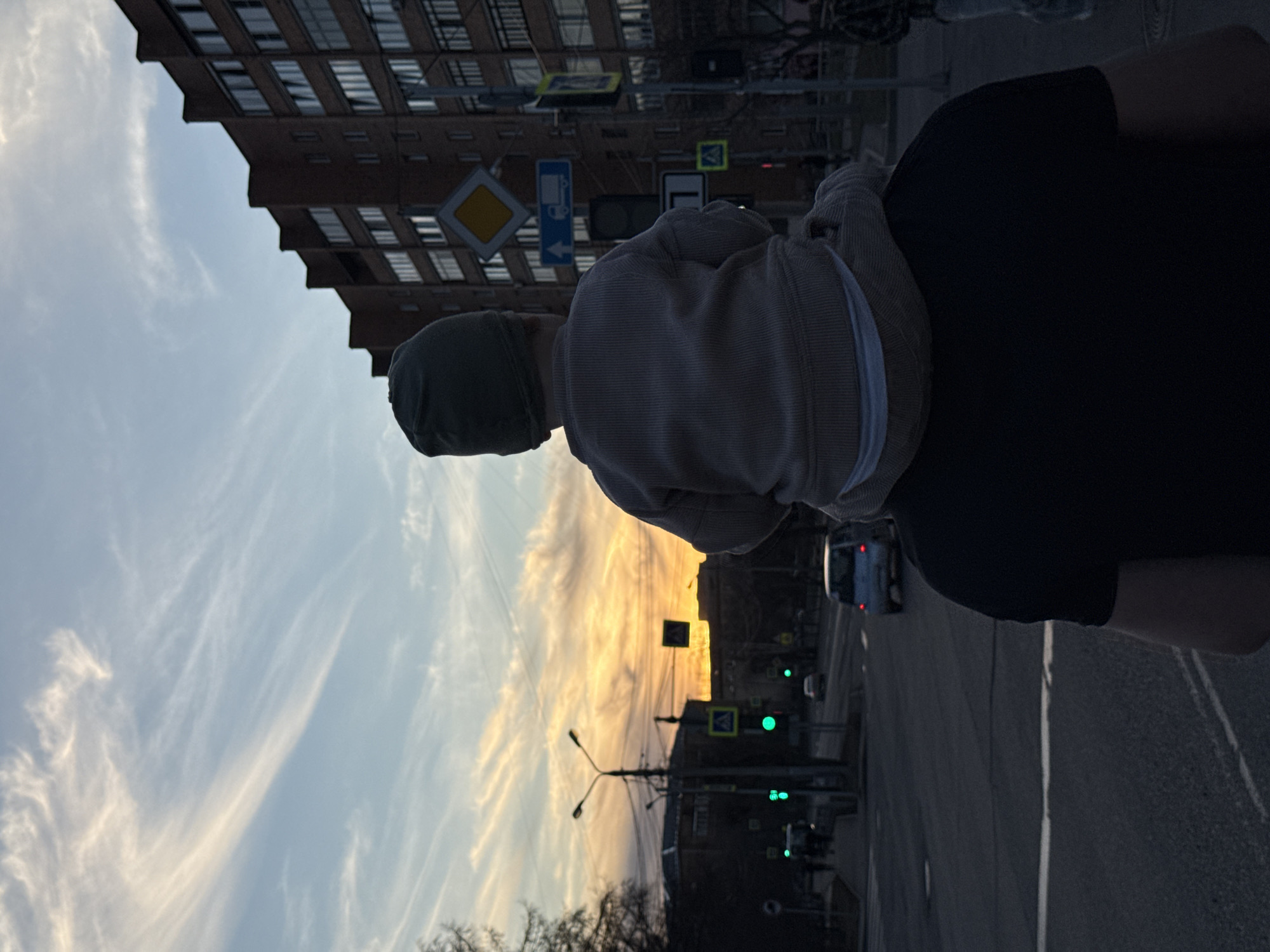 A medium shot captures a person holding a child in their arms against a city backdrop at dusk. The person, dressed in a dark-colored shirt, has the child positioned on their shoulder, obscuring much of their view. The child is wearing a gray hat and a striped gray top.
The background reveals an urban setting with a multi-story building to the left and street signs visible above. The sky is painted with hues of orange and blue, indicating either sunrise or sunset. Street lights and traffic signals line the street, adding to the city atmosphere. A vehicle with red lights is visible further down the street.
Based on the street signs and architectural style, the location appears to be Krasnoyarsk, Russia.