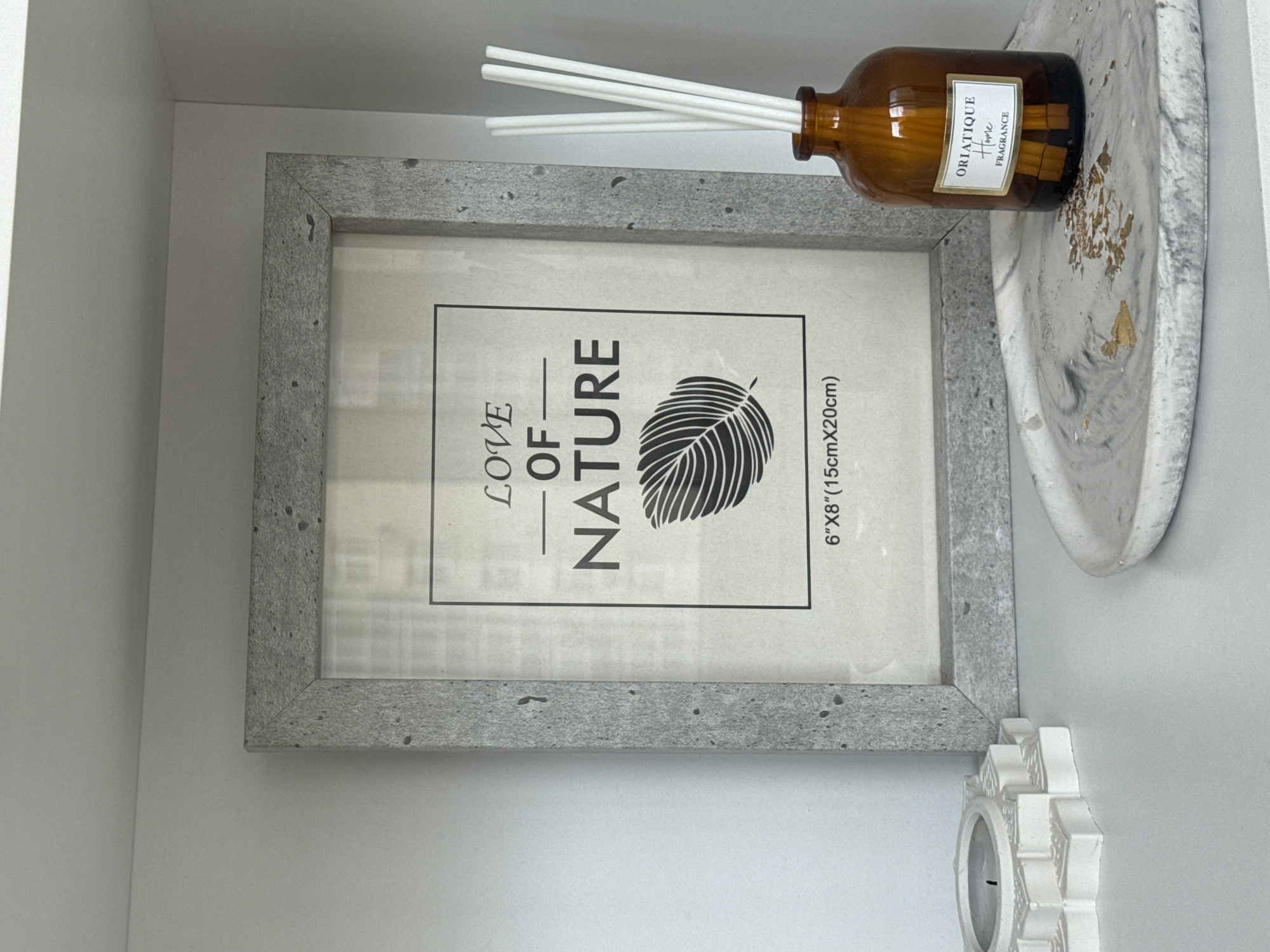 This is a close-up, eye-level, indoor shot of a shelf displaying decorative items. The shelf is white and the background is also white, suggesting an interior setting such as a home or a retail display. Prominently featured is a grey, textured photo frame containing a print with the words "LOVE OF NATURE" and a depiction of a leaf. To the right of the frame, there is a small amber glass bottle with a label that reads "ORIATIQUE Home FRAGRANCE," containing white diffuser reeds. In front of these items, on a marble-patterned tray, is a white, star-shaped candle holder with a partially burned candle. The scene is well-lit with soft, diffused light, casting no harsh shadows. There are no people or animals visible in the image, and no overt actions or interactions are occurring. The image does not provide any information that would allow for determining its location in Yekaterinburg, Russia.