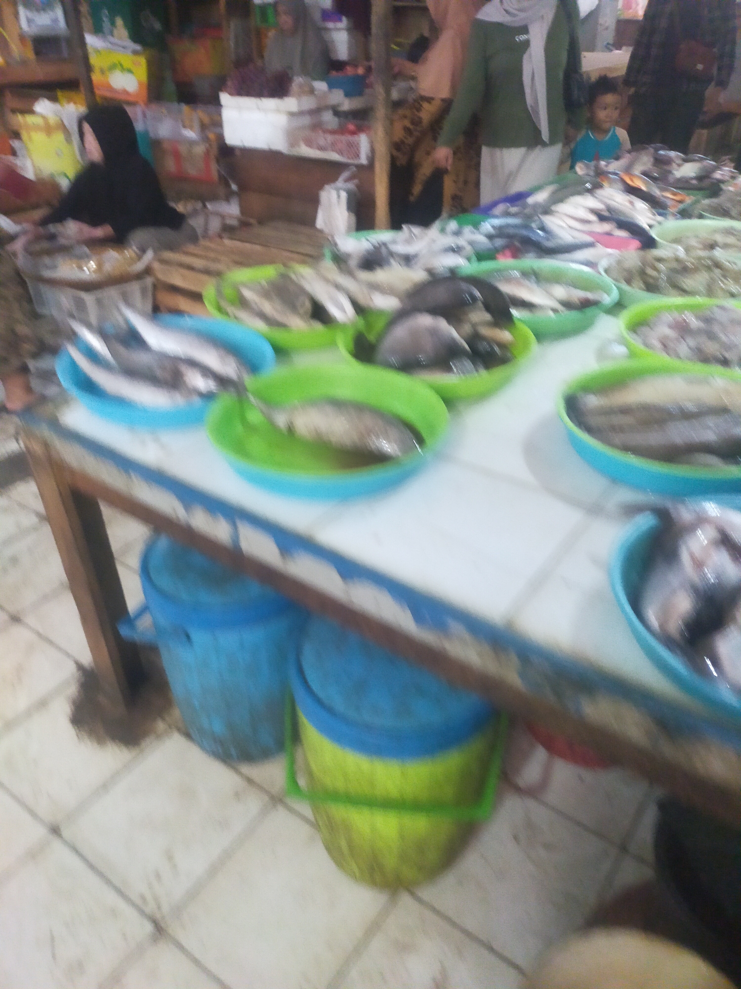 The image depicts a bustling fish market scene in Trayeman, Indonesia. In the foreground, several round green and blue containers are filled with fresh fish. The fish are of various sizes and types, some appearing whole and others filleted. A wooden table, likely a vendor's stall, is positioned in the middle ground, supporting the containers of fish. Behind the table, a woman wearing a black headscarf and a black garment is visible, seemingly engaged in preparing or displaying the fish. Further back, a child in a blue shirt stands near an adult wearing a green headscarf and a green shirt. The background reveals more market stalls with various goods, including what appears to be produce like grapes and other food items. The overall environment suggests a busy outdoor or semi-covered market, typical of local commerce in Indonesia. The lighting is somewhat dim, indicating it might be early morning or an indoor market with artificial lighting. The floor is tiled and appears somewhat wet, consistent with a market that sells fresh produce and seafood.