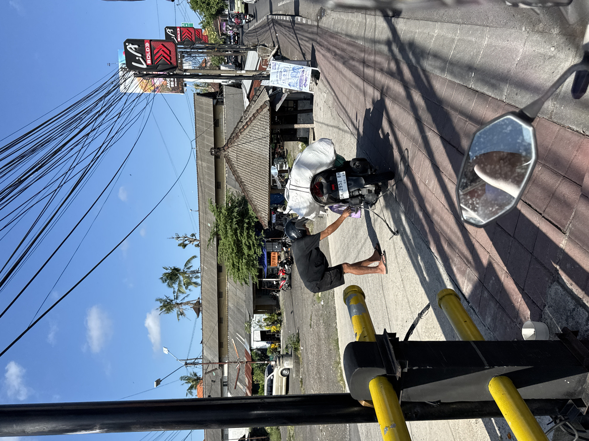 A man in a black t-shirt and shorts, wearing a black helmet, is standing on the sidewalk next to a parked motorcycle loaded with a large white bag. He is bending over, reaching towards the motorcycle, possibly securing the bag. The motorcycle has a license plate that reads "DK 2952 BH 06-26". The sidewalk is paved with red bricks and shows long, sharp shadows cast by overhead wires. To the left, a black pole with a yellow metal railing is visible. Behind the man and motorcycle, there are buildings with corrugated metal roofs, a small covered structure with a pitched roof, and several palm trees. In the background, a busy street with vehicles and signs, including a "LA BOLD" billboard, indicates an urban environment. The sky is blue with scattered white clouds, suggesting a sunny day. The scene appears to be in North Kuta, Indonesia, as indicated by the overall setting and streetscape.