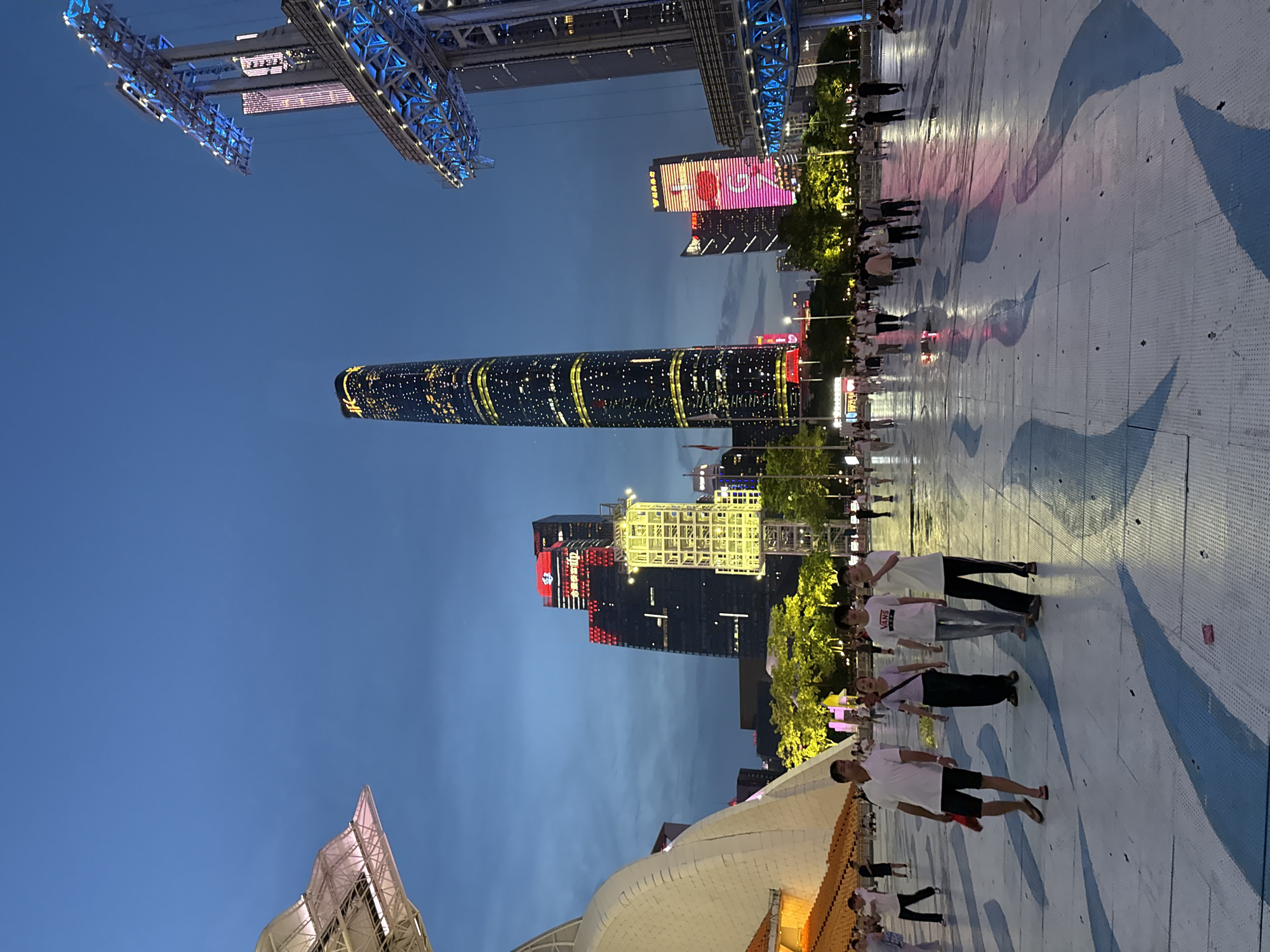 This is a medium shot of a public plaza at dusk, possibly in Tianhe District, China, featuring a mix of modern architecture and people. In the background, a very tall, slender skyscraper with illuminated yellow and white bands wraps around its facade, likely the Canton Tower. To the left of this skyscraper is a shorter, dark building with prominent red and white illuminated signage. To the right, a partially visible, large, modern structure with a white, curved roof and visible supporting beams is present. Further to the right, a tall, blue illuminated structure with multiple levels of lights and platforms dominates the view. Several other buildings of varying heights and illumination are scattered throughout the background.
The plaza itself is paved with large, light-colored tiles, some of which have blue, wave-like patterns. The surface appears wet in places, reflecting the lights from the surrounding buildings. A group of people are walking across the plaza, most of them in casual attire. On the left side of the foreground, a man in a white polo shirt and black shorts walks towards the viewer. To his right, a woman in a dark top and pants walks in the same direction. Next to her are two young men, one in a white t-shirt with "VANS" printed on it, and the other in a white t-shirt and glasses. Further into the plaza, more people are seen strolling, some in groups. Trees with yellow lights are visible between the plaza and the buildings. The sky is a deep blue, indicating the transition from day to night, with some wispy clouds. The overall impression is an active, urban evening scene.