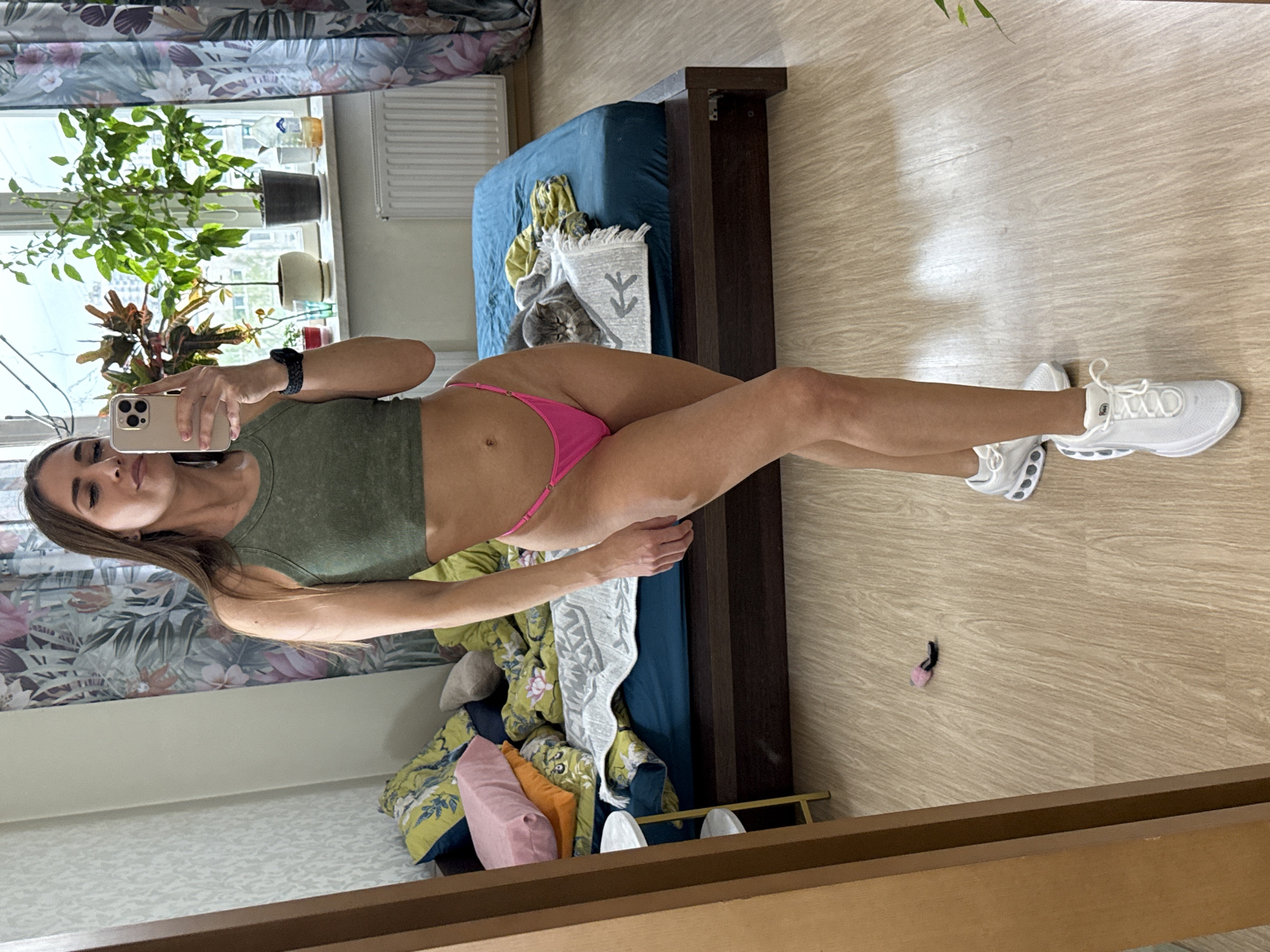 A woman in a green top and pink underwear is taking a selfie in a room. She is standing near a bed with blue sheets and colorful pillows. There is a cat on the bed. The floor is wooden. A plant is on the windowsill. A radiator is also present near the window. The image appears to be taken in a mirror. The Saint Petersburg, Russia location cannot be determined.