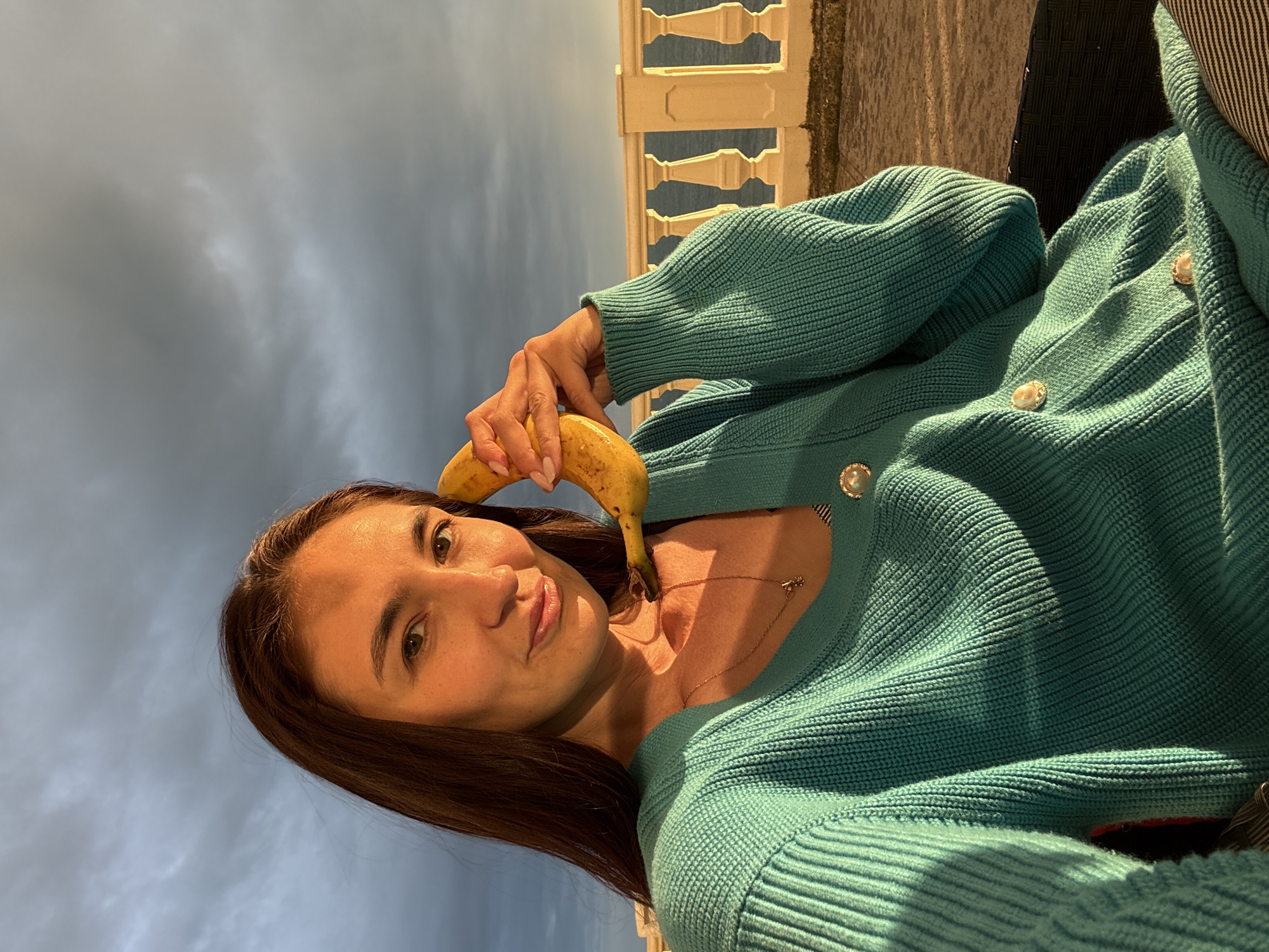 A young woman holds a banana near her ear, posing for a selfie outdoors in Sochi, Russia. She is wearing a teal-colored knitted sweater with pearlescent buttons, and a gold necklace. The sky behind her is partly cloudy. In the background, there is a white balustrade and vegetation.