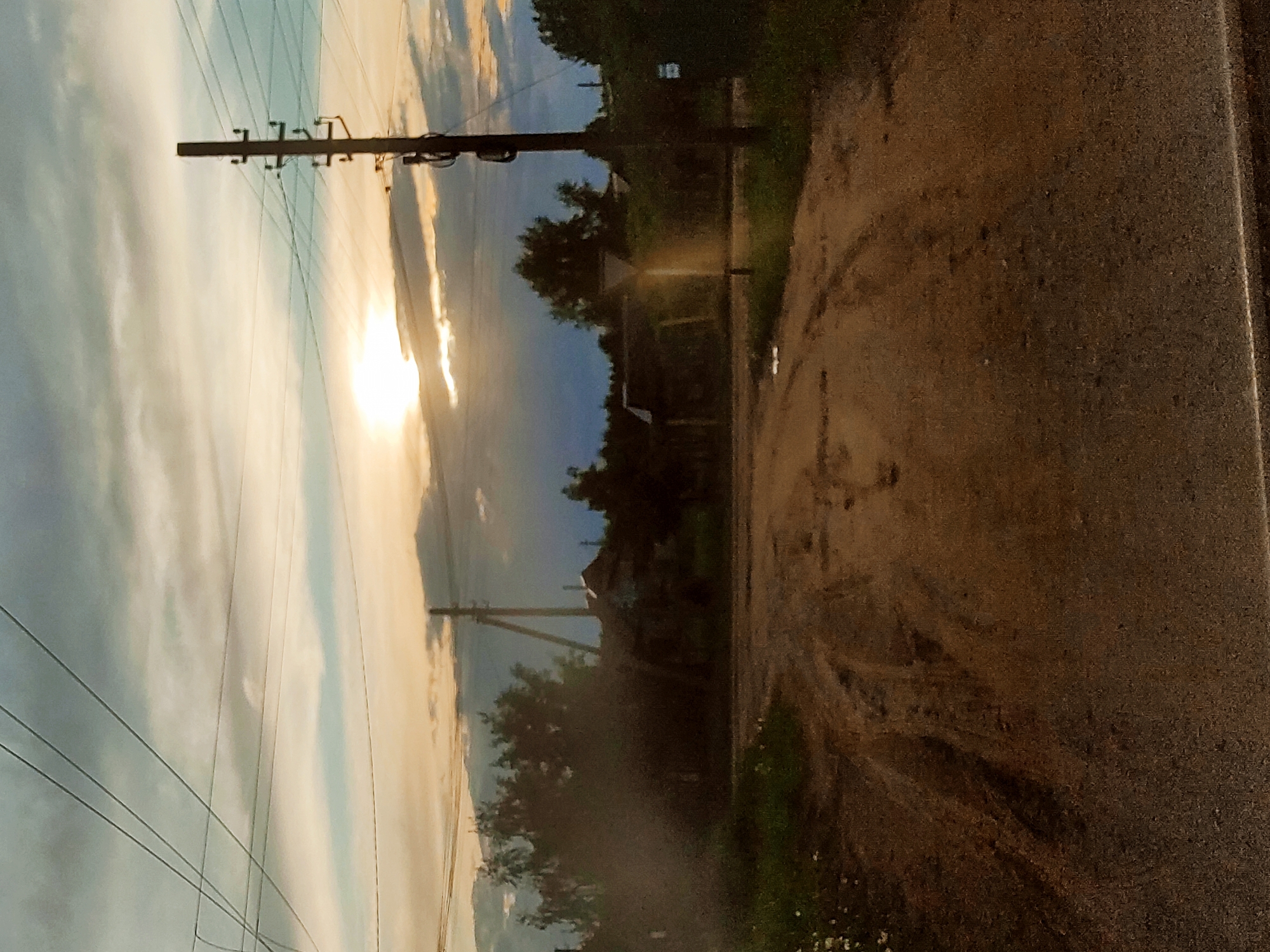 The image shows a rural road scene at sunset in Ольховка, Russia. The road, possibly dirt or gravel, is visible on the right with tire tracks. On the left, utility poles with power lines stretch across the sky, partially obscuring the setting sun. Trees and a few buildings are visible in the middle ground. The overall lighting indicates it is either early morning or late evening.