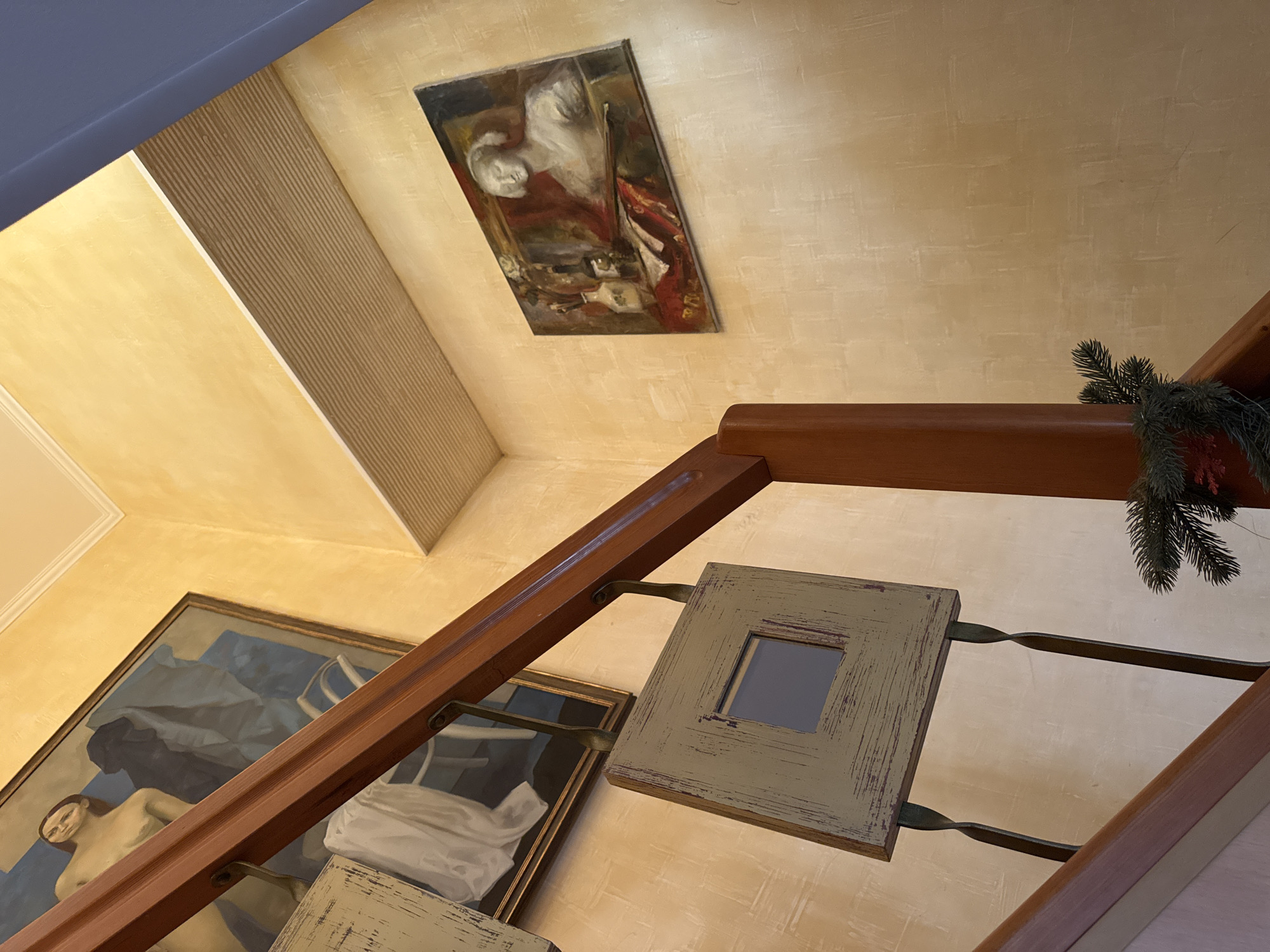 The image captures an interior view of a staircase with artworks displayed on a yellow-textured wall. A wooden banister runs diagonally across the frame. To the left, a large framed painting depicts a nude woman and a dark, abstract form. Below this, a square decorative frame with a reflective surface hangs from the banister. To the right, another framed oil painting features a bust of a woman with what appears to be musical instruments and a vase on a table in the foreground. A strand of evergreen garland with a red ornament is visible near the bottom right of the frame, suggesting a festive decoration. The lighting suggests an indoor setting, possibly a home or a public space with an artistic ambiance. There is no visible indication that this scene is located in Yeysk, Russia.