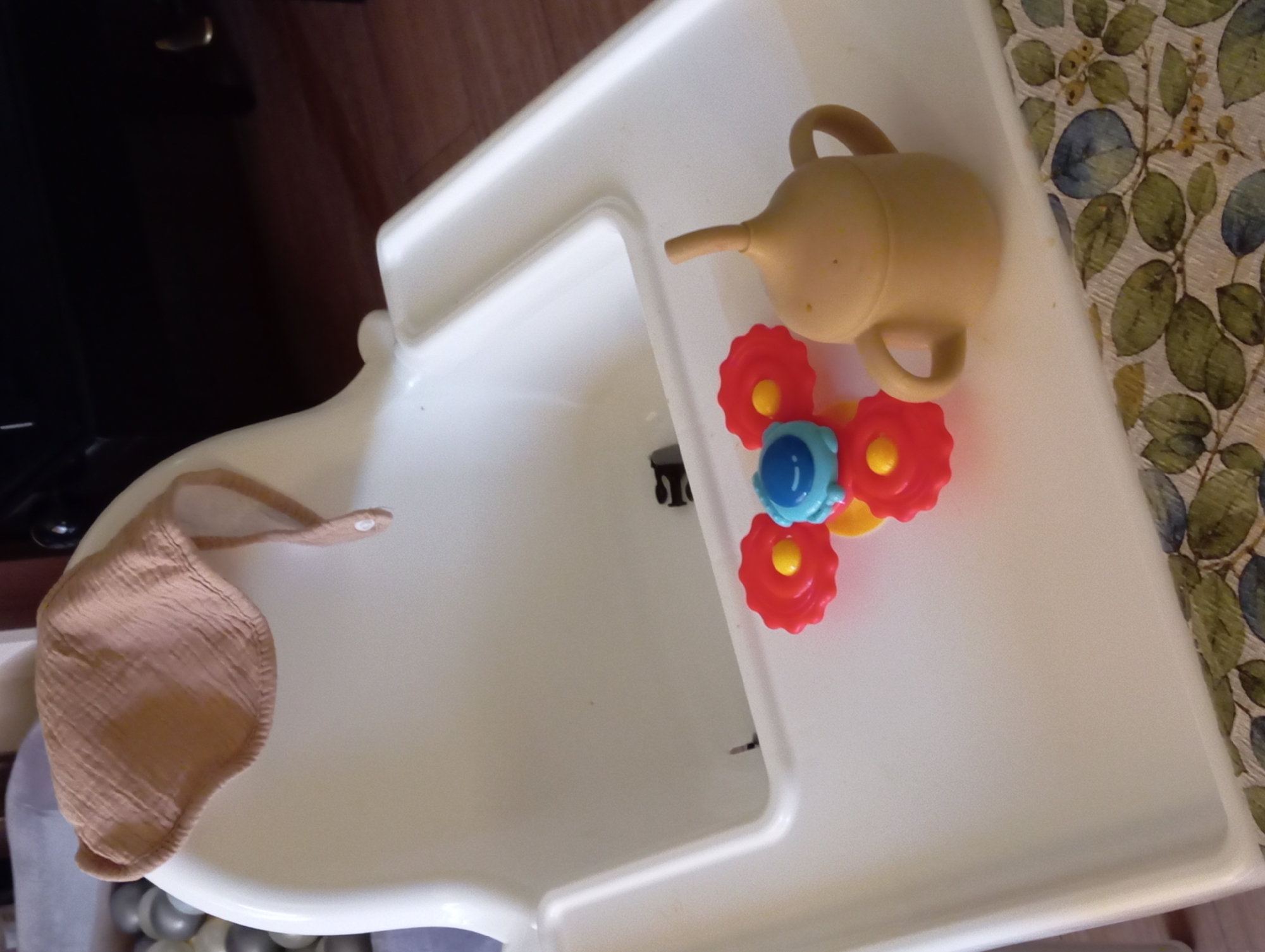 A high-angle medium shot captures a white plastic baby high chair with a bib draped over the top. On the tray of the high chair, there are three colorful plastic toys: a red and yellow flower-shaped toy, a blue and yellow toy, and a beige sippy cup. The background of the image is blurred, showing a dark wooden floor and a dark piece of furniture. The scene is indoors and appears to be in a home environment, possibly a kitchen or dining area. There is no indication of the location being in Baden, Austria.