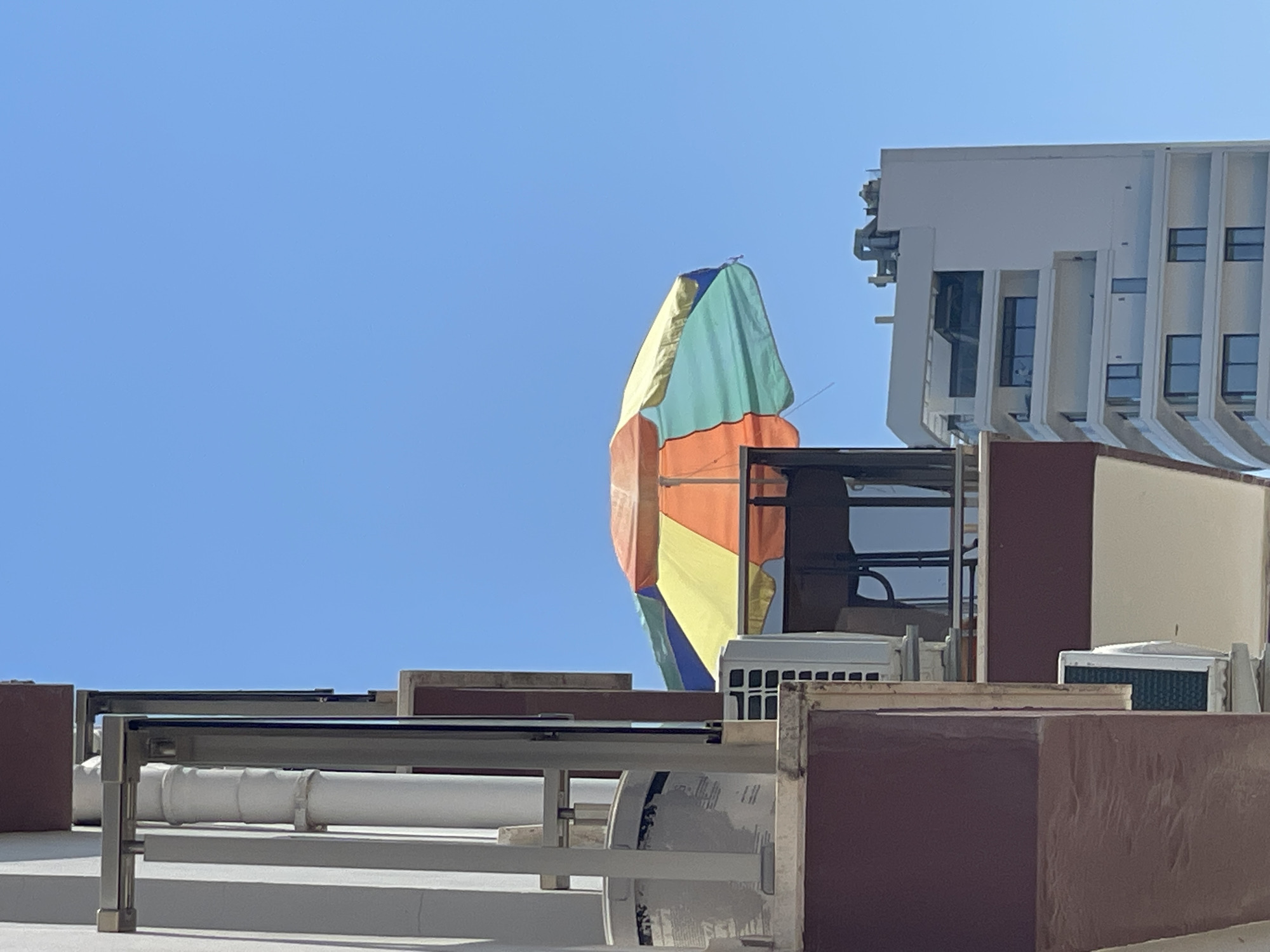 The image depicts a bright, sunny day with a clear blue sky. In the foreground, a section of a building is visible on the left, featuring maroon and white exterior walls, a silver pipe, and what appears to be an air conditioning unit. To the right, a colorful umbrella, with segments of orange, yellow, green, and blue, is open and tilted. Behind the umbrella, a modern white high-rise building with balconies and large windows is visible. The scene suggests an urban or resort setting, possibly in Trikomo, Northern Cyprus, given the architectural style and the presence of a beach umbrella. There are no people visible in the frame.