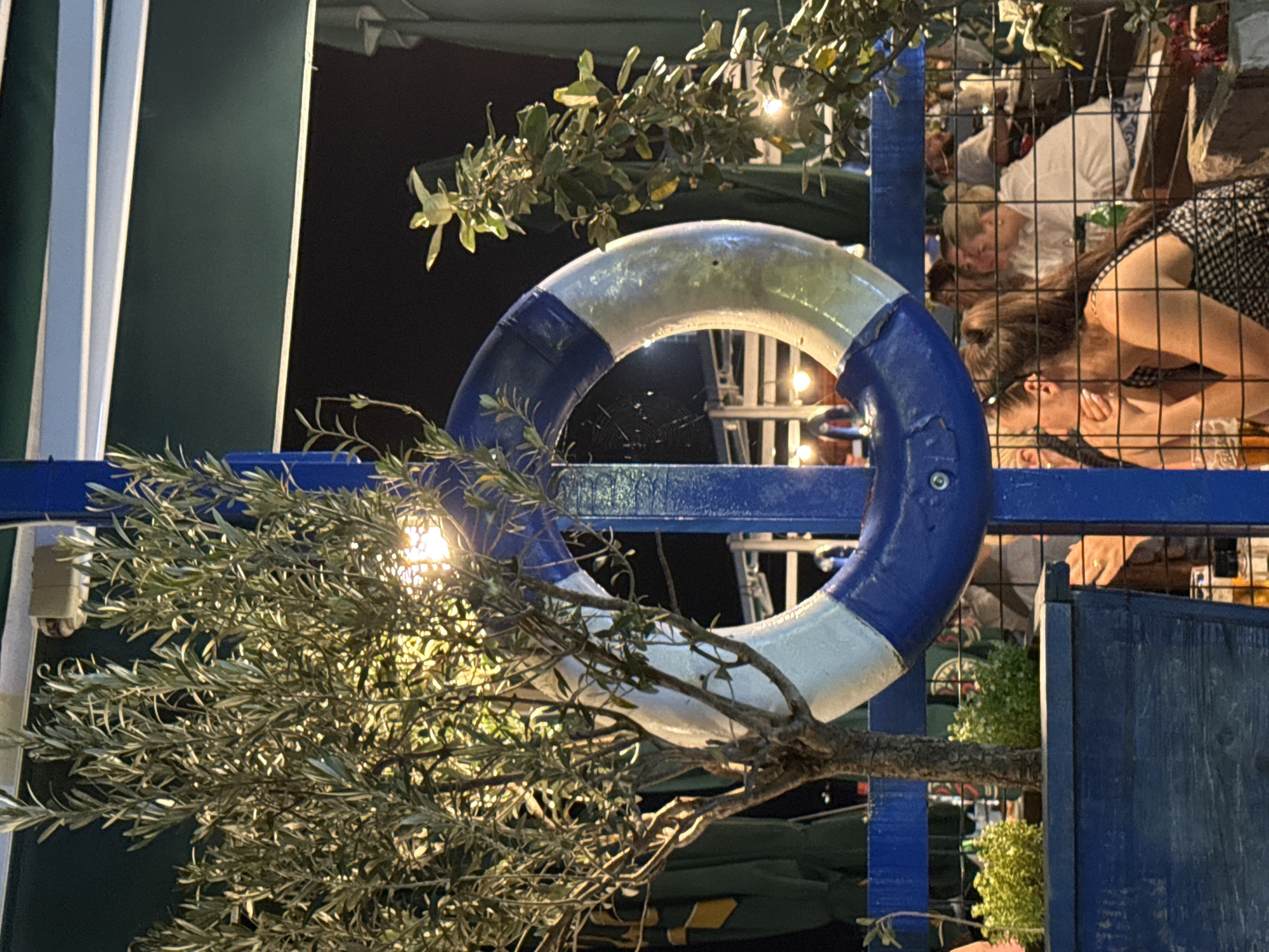 The image depicts a nighttime outdoor scene at a restaurant in Budva, Montenegro. A blue and white life preserver is prominently displayed, suspended from a blue pole. To the left of the life preserver is a small olive tree in a blue wooden planter. Behind the life preserver and olive tree, patrons are visible seated at tables, some obscured by a wire mesh fence. The lighting suggests a casual dining atmosphere. The overall impression is that of a coastal restaurant or bar.