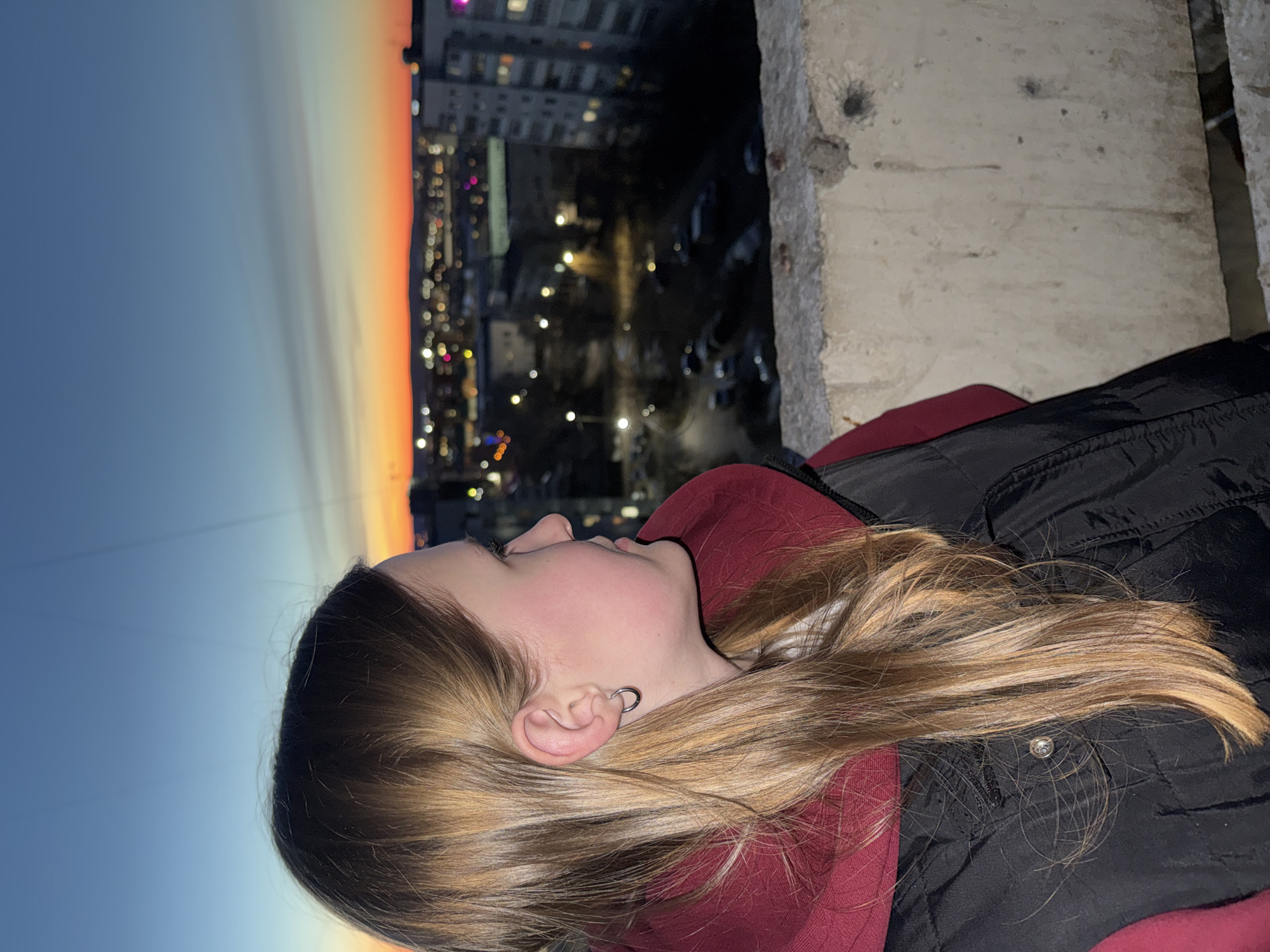 A young woman is on a balcony at dusk or night. She is facing away from the camera looking at the buildings of Chaikovsky, Russia below. She is wearing a dark jacket and a maroon hoodie. The balcony is concrete and there is a sky of blue, yellow, and orange over the city.