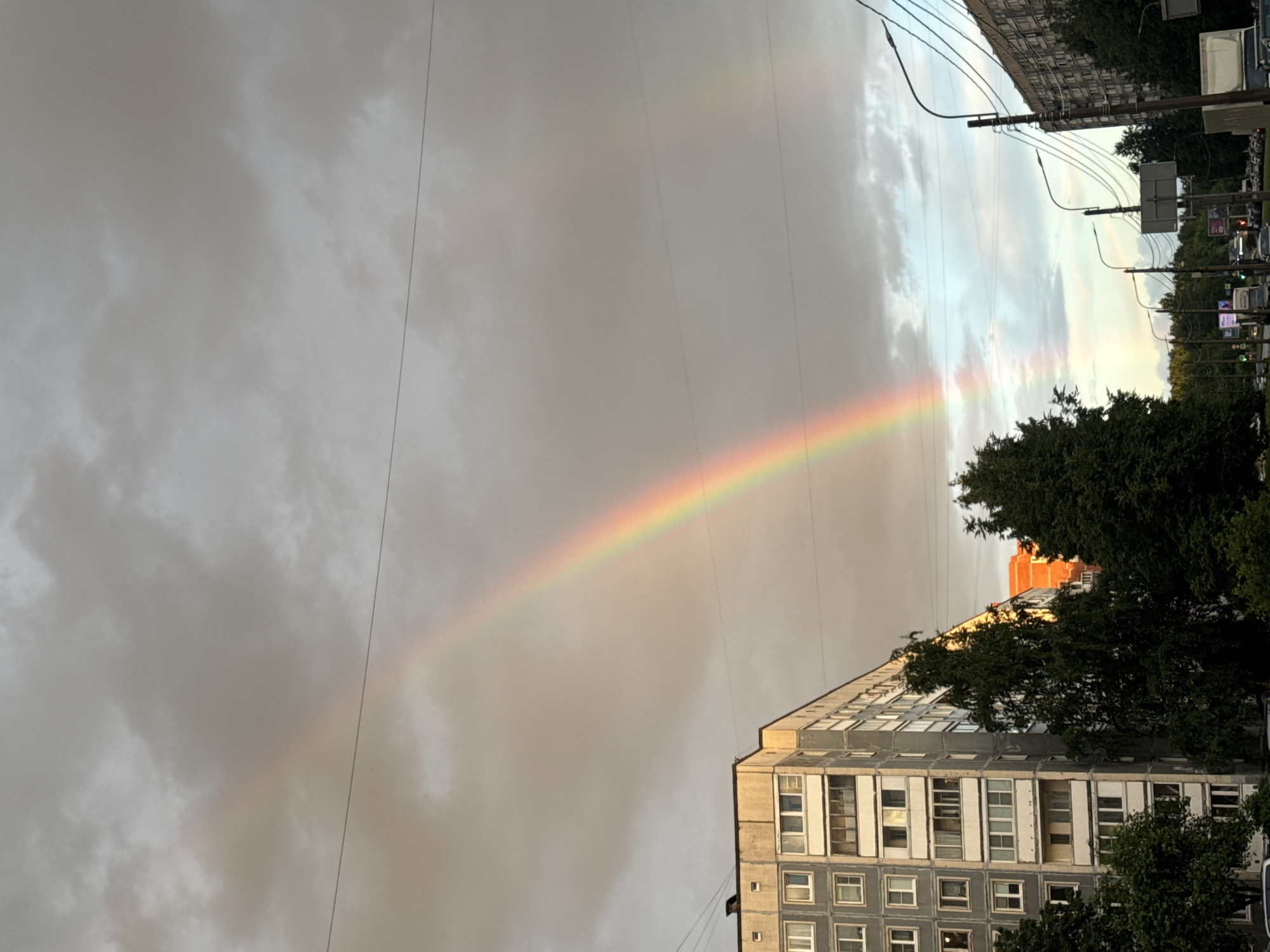 A rainbow arcs across a cloudy sky above a street scene in an urban environment. The primary rainbow, vibrant with red, orange, yellow, green, blue, and violet hues, begins in the upper left and extends diagonally downwards to the right. A fainter, secondary rainbow is visible to the right of the primary one.
Dominating the left side of the frame is a multi-story apartment building with a gray facade and numerous windows. Dense green trees are positioned in front of the building and further down the street. To the right, another apartment building, also appearing to be residential, runs along the side of the street.
The street itself is lined with utility poles and streetlights, and several vehicles are visible, including cars and what appears to be a truck. The sky is filled with overcast, gray clouds, with some lighter patches suggesting the sun might be breaking through in places, contributing to the rainbow phenomenon. The overall impression is a somewhat muted urban landscape dramatically illuminated by the natural spectacle of a double rainbow.