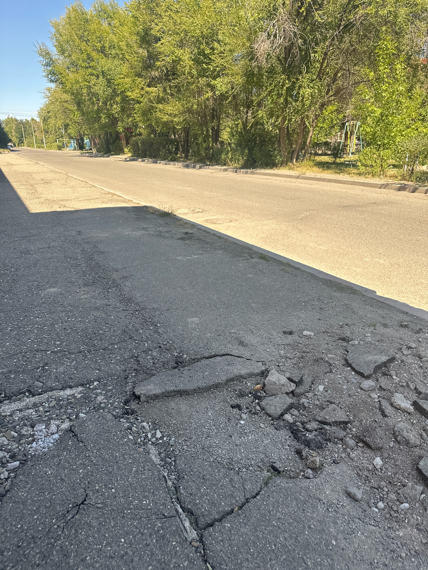 The image shows a street scene in Gyumri, Armenia. A paved road runs horizontally across the frame, lined with mature trees on the right side. The road is flanked by a cracked and crumbling asphalt sidewalk in the foreground. The sidewalk shows significant damage, with large pieces of asphalt broken and scattered, revealing loose gravel and debris underneath. The sun casts a strong shadow across the left side of the foreground, indicating a bright, sunny day. In the distance, the road recedes and appears to be in better condition, with a few cars visible. The overall impression is one of urban infrastructure in disrepair, juxtaposed with natural elements.