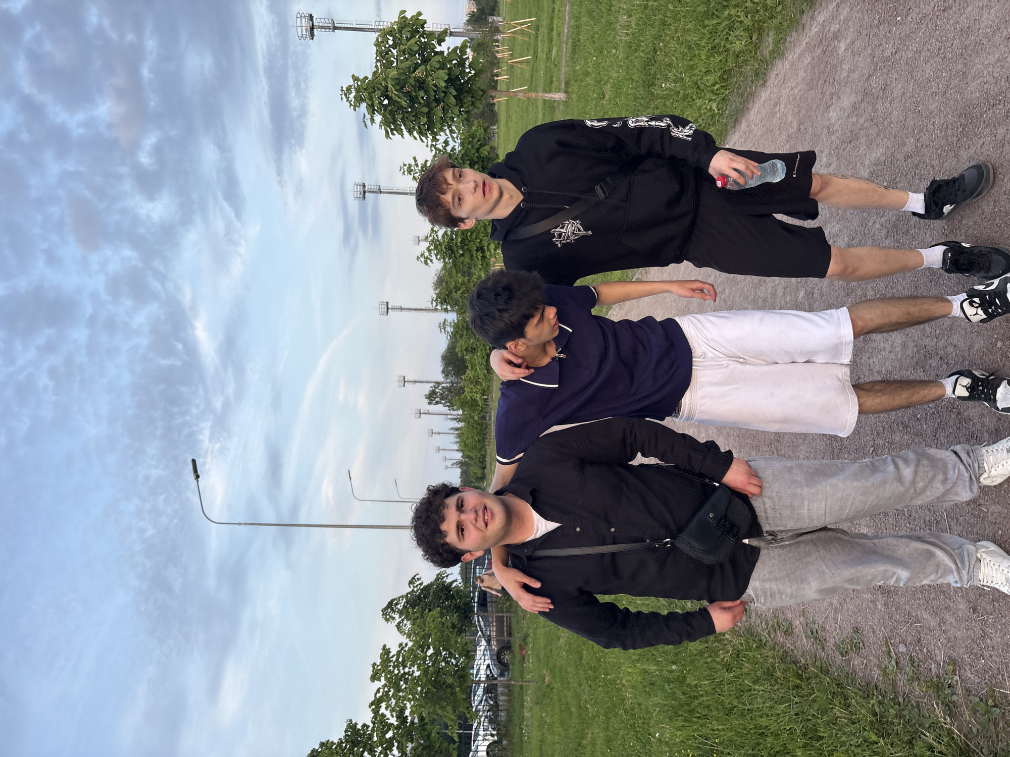 Three young men are standing on a gravel path in a park. The man on the left has curly dark hair and is wearing a black button-up shirt, grey pants, and white sneakers. He has a black crossbody bag. The man in the middle has dark hair and is wearing a navy blue polo shirt with white trim, white shorts, and black and white sneakers. The man on the right has light brown hair and is wearing a black hoodie, black shorts, white socks, and black sneakers. He is holding a water bottle. In the background, there are trees, streetlights, and a parking lot with cars. The sky is cloudy. The location is the Sinyavino park in Sinyavino, Russia.