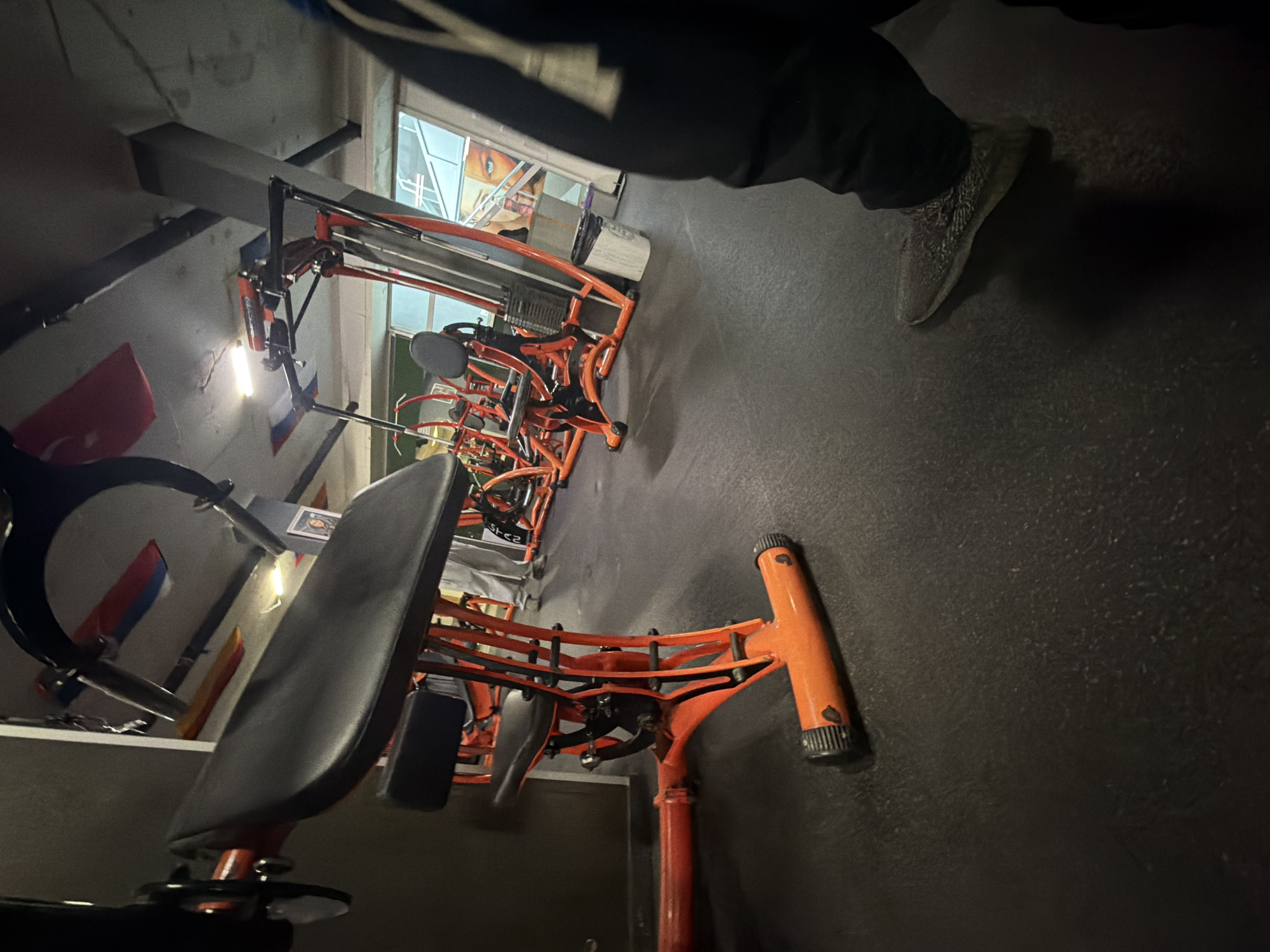 The image shows a fitness center in Burgas, Bulgaria. In the foreground is a weight machine with an orange frame and a black seat. Several similar machines are lined up behind it. The floor is a dark gray material. In the upper right corner of the image, a person wearing gray sneakers and black pants is visible. Flags of different nations are displayed on the wall.