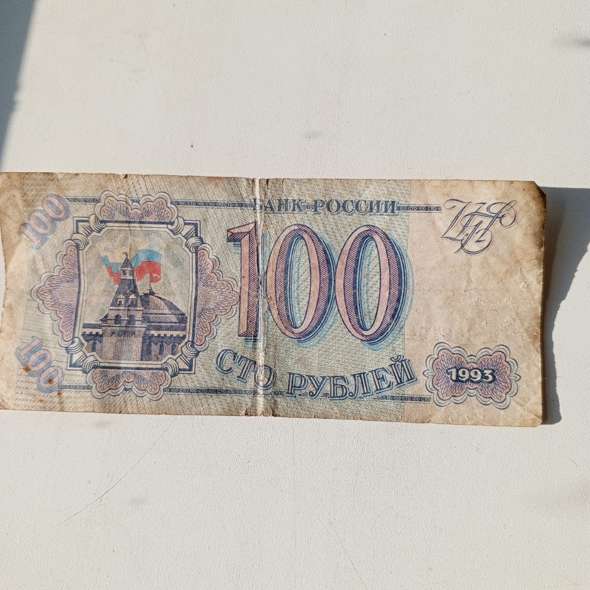 The image shows a faded, worn 100 Russian Ruble banknote from 1993. The banknote features a depiction of the Spasskaya Tower of the Moscow Kremlin, with Russian flags flying from its spires. The words "БАНК РОССИИ" (Bank of Russia) are printed across the top, and "СТО РУБЛЕЙ" (One Hundred Rubles) is printed at the bottom. The number "100" is prominently displayed in the top left and center of the note, and the year "1993" is visible in the bottom right. The banknote is lying on a white, textured surface, likely a table or countertop. There is a shadow cast on the surface from the left side of the image, suggesting a light source from that direction. The banknote appears to be creased and slightly torn along the edges, indicating age and use. There are no people or any other objects visible in the image. The image does not provide any information about the location in the city of Sayanogorsk, Russia.
