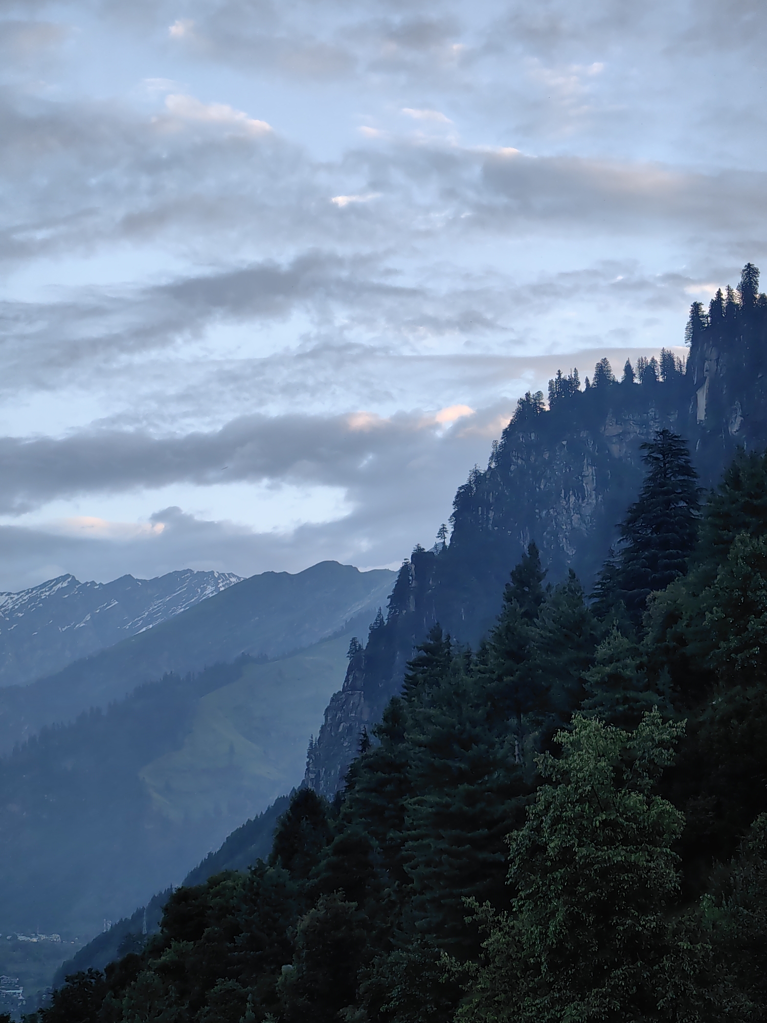 This image depicts a mountainous landscape with a dramatic, steep rock face covered in dark green coniferous trees dominating the foreground and right side. The trees are densely packed and vary in height, with some taller specimens reaching towards the sky. Beyond this prominent rock face, further mountain ranges recede into the distance, appearing in shades of blue and grey, with some of the higher peaks showing patches of snow. The sky above is filled with a mixture of light and dark grey clouds, with hints of pale pink and yellow suggesting either dawn or dusk. In the lower left portion of the image, a small village or settlement is faintly visible nestled in the valley below the mountains, with buildings appearing as small clusters of light and dark shapes. The overall atmosphere is serene and somewhat dramatic, characterized by the ruggedness of the mountains and the subdued lighting. This scene is likely located in the vicinity of Vashisht, India, a region known for its mountainous terrain and natural beauty.
