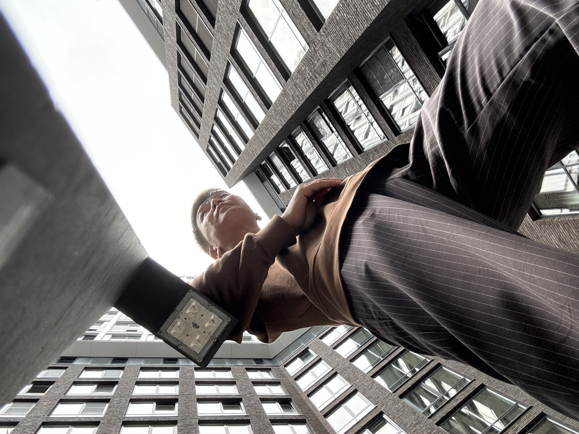 The media file captures a low-angle shot looking up towards a man in striped pants and a brown top, standing in front of a modern, dark brick building with many windows. The building appears to be an apartment or office complex, with a grey, contemporary architectural style. The perspective emphasizes the height of the building and the man's position relative to it.
In the foreground, a concrete structure with a built-in light fixture extends into the frame, partially obscuring the view of the man and the building. The light fixture has a square face with visible LEDs. The man is looking upwards, with his hand resting on his chest, suggesting a moment of reflection or observation. His expression is neutral.
The time of day appears to be daytime, with bright, overcast skies contributing to the even lighting of the scene. There are no specific weather indicators, but the general brightness suggests it is not raining or excessively dark. The setting is urban, with the towering architecture of Moscow, Russia, being the dominant visual element. The modern design of the buildings, including the windows and brickwork, are characteristic of contemporary city construction.
No text is visible in the image. The overall impression is one of urban exploration or a candid moment captured in a metropolitan environment.