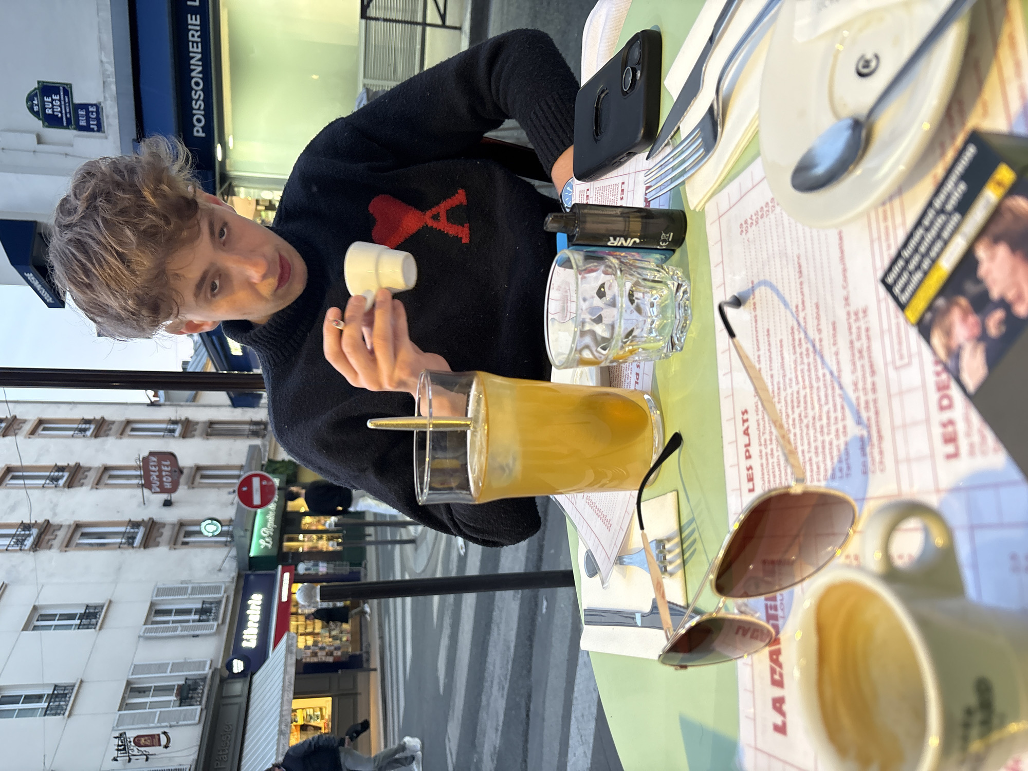 An overhead-angle, candid photograph depicts a young male seated at an outdoor cafe table in Paris, France, specifically on Rue Juge in the 15th Arrondissement, identifiable by a visible street sign and the "DUPLEIX HOTEL" sign in the background. The male, with short, curly light brown hair, wears a black turtleneck sweater featuring a red heart-shaped logo (AMI Paris). He holds a small white espresso cup in his right hand, with a lit cigarette held between his index and middle fingers of the same hand. His gaze is directed downwards towards the cup.
The light green cafe table is set with a white placemat, an open menu, and various items. These include a tall clear glass containing an amber beverage with ice and a gold stirring spoon, an empty clear glass, a pair of aviator-style sunglasses with gold frames, a black cylindrical vape pen labeled "JNR", and a black smartphone in a black case lying face down. Also present are a small white plate with a silver spoon, and a wrapped set of cutlery (fork and knife). A cigarette pack, displaying French health warnings and an image of two faces, is partially visible at the bottom right.
The background reveals a bustling urban street scene during what appears to be dusk or an overcast day. Multi-story buildings with light-colored facades and numerous windows, some with white shutters, line the street. Ground-floor businesses include a "Librairie" (bookstore), a "Poissonnerie" (fishmonger), and a "Pâtissier" (pastry shop). A red circular "no entry" sign is prominent. Several pedestrians are visible walking on the street, including one near a zebra crossing, and another near a storefront.