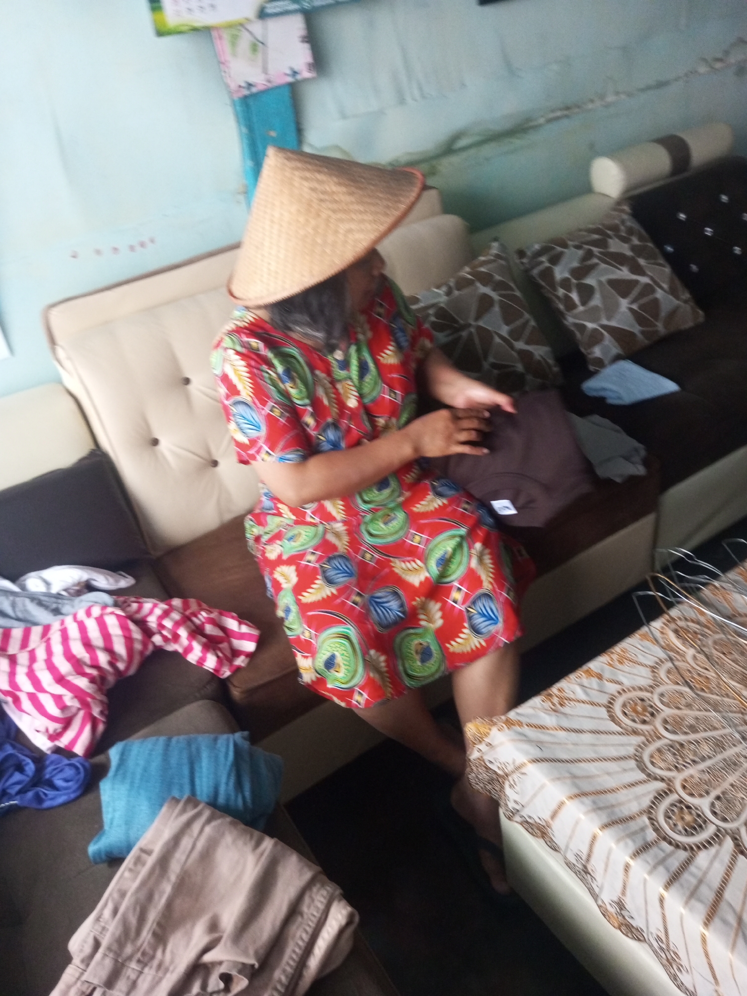 The image shows a woman sitting on a sofa, wearing a conical hat and a red dress with a floral pattern. She is holding a folded brown garment in her hands. The sofa is upholstered in beige and brown fabric, and there are several decorative pillows on it. In the foreground, there are piles of folded clothes, including a striped pink and white shirt, a blue towel, and a pair of beige pants. A table covered with a decorative cloth is also visible. The wall behind the sofa is painted light blue and has some framed pictures or posters. The scene appears to be an indoor setting, possibly a living room, in a residential area of Andongsili, Indonesia.