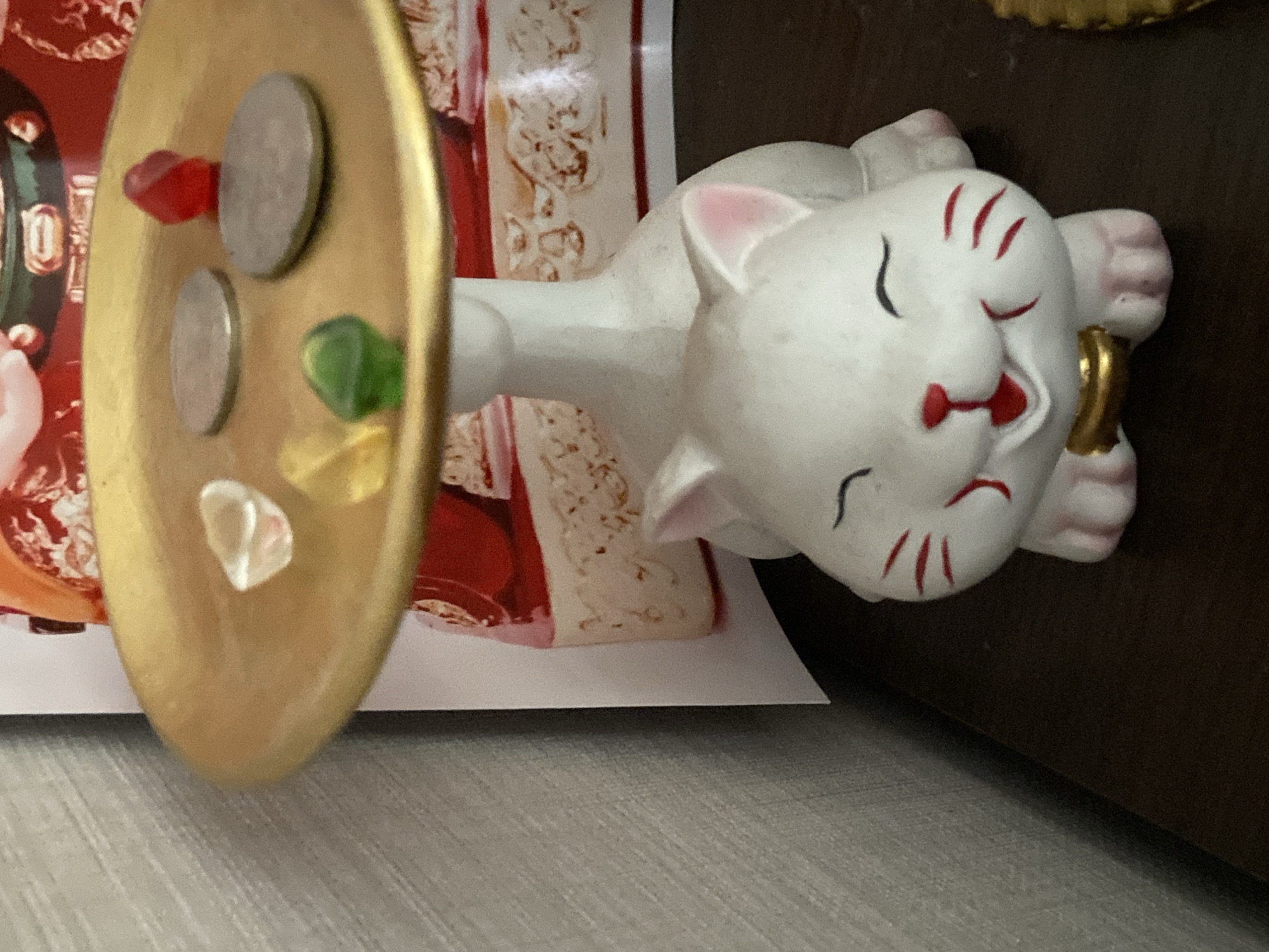 The image shows a decorative white ceramic cat figurine with red accents, sitting on a dark surface. The cat has its eyes closed in a smile and is wearing a gold collar. A gold-colored tray rests on a white stand behind the cat. The tray holds two silver coins and three small, colorful, translucent objects that appear to be beads or gems (one red, one green, and one clear or yellow). In the background, a portion of a colorful, possibly decorative, object with red and white elements is visible. The lighting suggests an indoor setting. There is no information in the image that allows for the identification of the city of Ulan-Ude, Russia.
