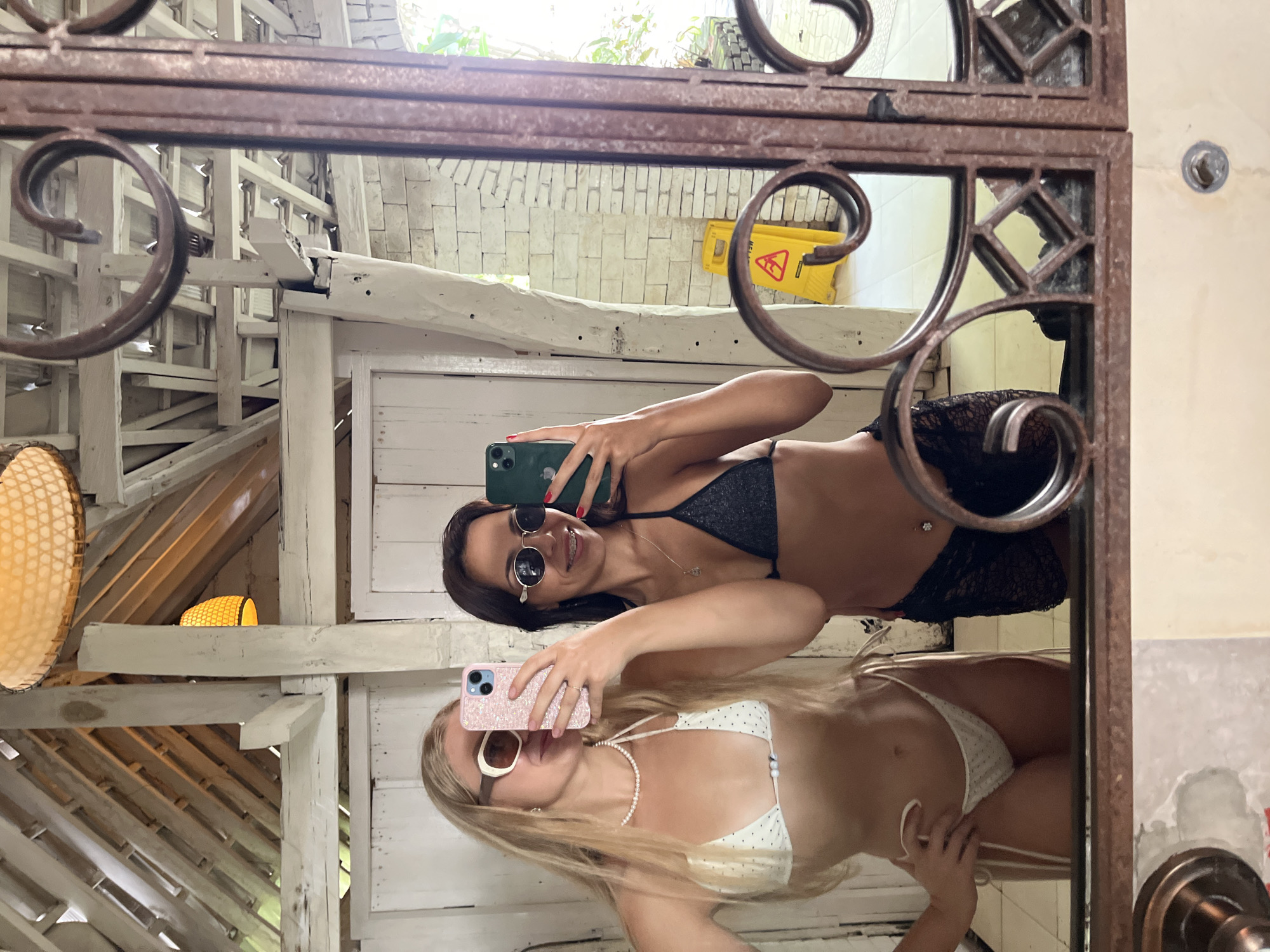 An image, oriented horizontally, depicts two young women taking a mirror selfie. The mirror itself features an ornate, dark brown, wrought-iron style frame with intricate scrollwork.
The woman closer to the camera has blonde hair, wears a white polka-dot string bikini with a matching bottom, a pearl necklace, and white-framed sunglasses. She is holding a light pink smartphone with a glittery case, positioned to capture the reflection. She is smiling subtly.
Behind her, the second woman has dark hair, wears a dark grey or black shimmering bikini top, a black lace-patterned bottom, and round dark sunglasses. She holds a dark green smartphone, also angled for the selfie, and sports a broad smile. A small silver necklace and what appears to be a belly button piercing are visible.
The background, visible as a reflection in the mirror, reveals a rustic, open-air structure. It features white-painted wooden beams and slats, some appearing weathered. Elements of a tropical setting are present, including a woven, circular light fixture and another smaller, basket-weave light source. In the upper right corner of the reflection, a bright yellow "WET FLOOR" sign with a black warning symbol is visible. Behind this sign, a light-colored brick or stone wall can be seen, along with some green foliage. The scene suggests a bright, daytime setting. Both women appear relaxed and happy.