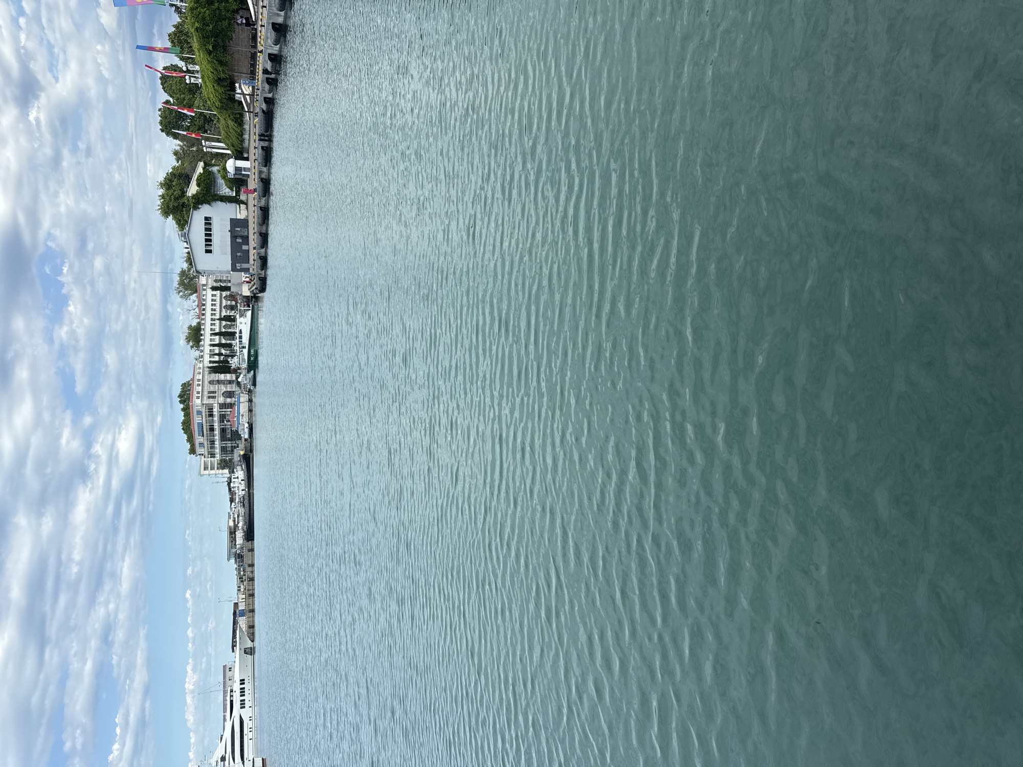 This is a photograph of the Sochi Sea Port in Sochi, Russia, on a cloudy day. The foreground is dominated by the turquoise water of the harbor, with gentle waves creating a textured surface. In the middle ground, a long pier extends into the harbor, lined with black fenders. Several boats and yachts are docked along the pier. A large white yacht is prominently visible on the left side of the image. In the background, a white building with columns and arched windows, characteristic of Stalinist architecture, stands along the waterfront. To the right of this building, a modern white building with a flat roof and large windows is situated. Flags of various nations fly from poles near the buildings. Green trees and foliage are visible behind the structures, and the sky above is filled with scattered white clouds. There are a few people visible on the pier in the distance.