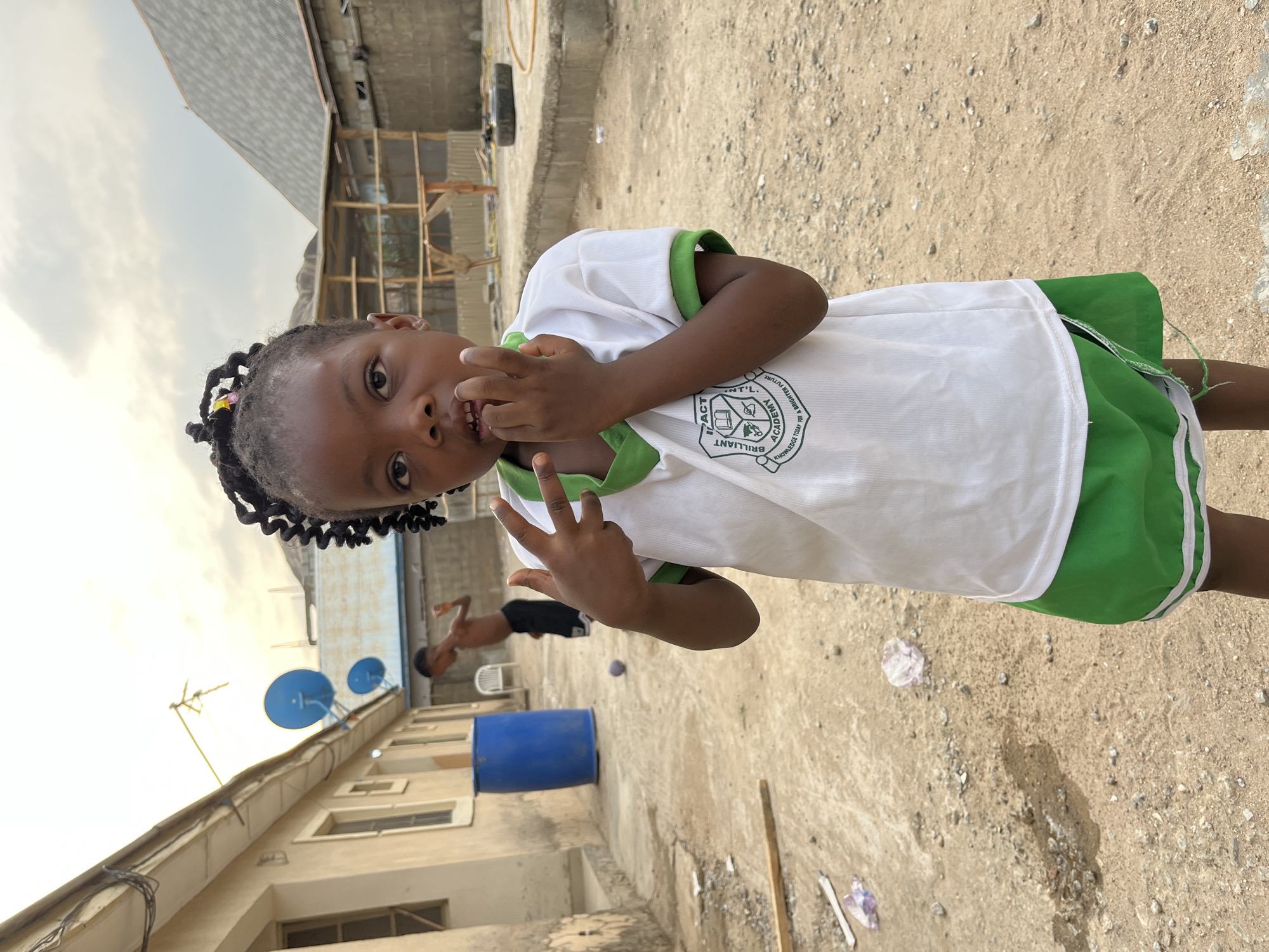 A young child stands in a dirt-covered area in Madalla, Nigeria. She is wearing a white shirt with green trim and green shorts. The shirt has a logo on it that says "Impact Brilliant Academy". She is holding her fingers up to her mouth, appearing to make a gesture. In the background, there is a building with blue satellite dishes and a blue barrel. Another person is faintly visible in the background, engaging in some activity. The environment is typical of a residential area, with buildings of mixed construction and a dusty ground. The sky is overcast.