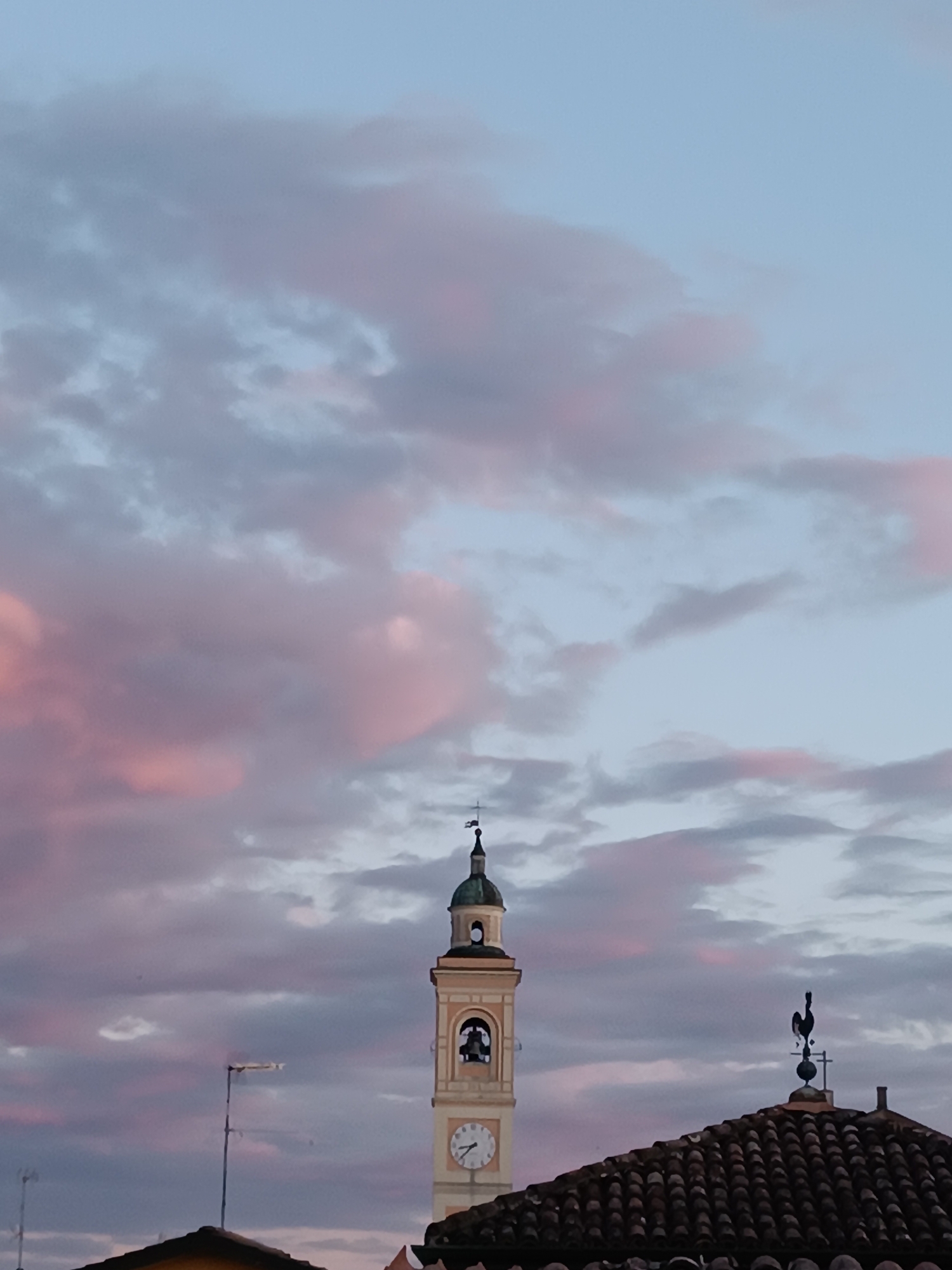 The image shows the church tower of the San Nicola di Bari Parish Church in Viadana, Italy, against a backdrop of pink-tinged clouds at dusk. The tower is beige and has a clock face and a green dome with a spire. To the right, a roof with terracotta tiles is visible, topped with a rooster weather vane. An antenna is visible in the lower left.