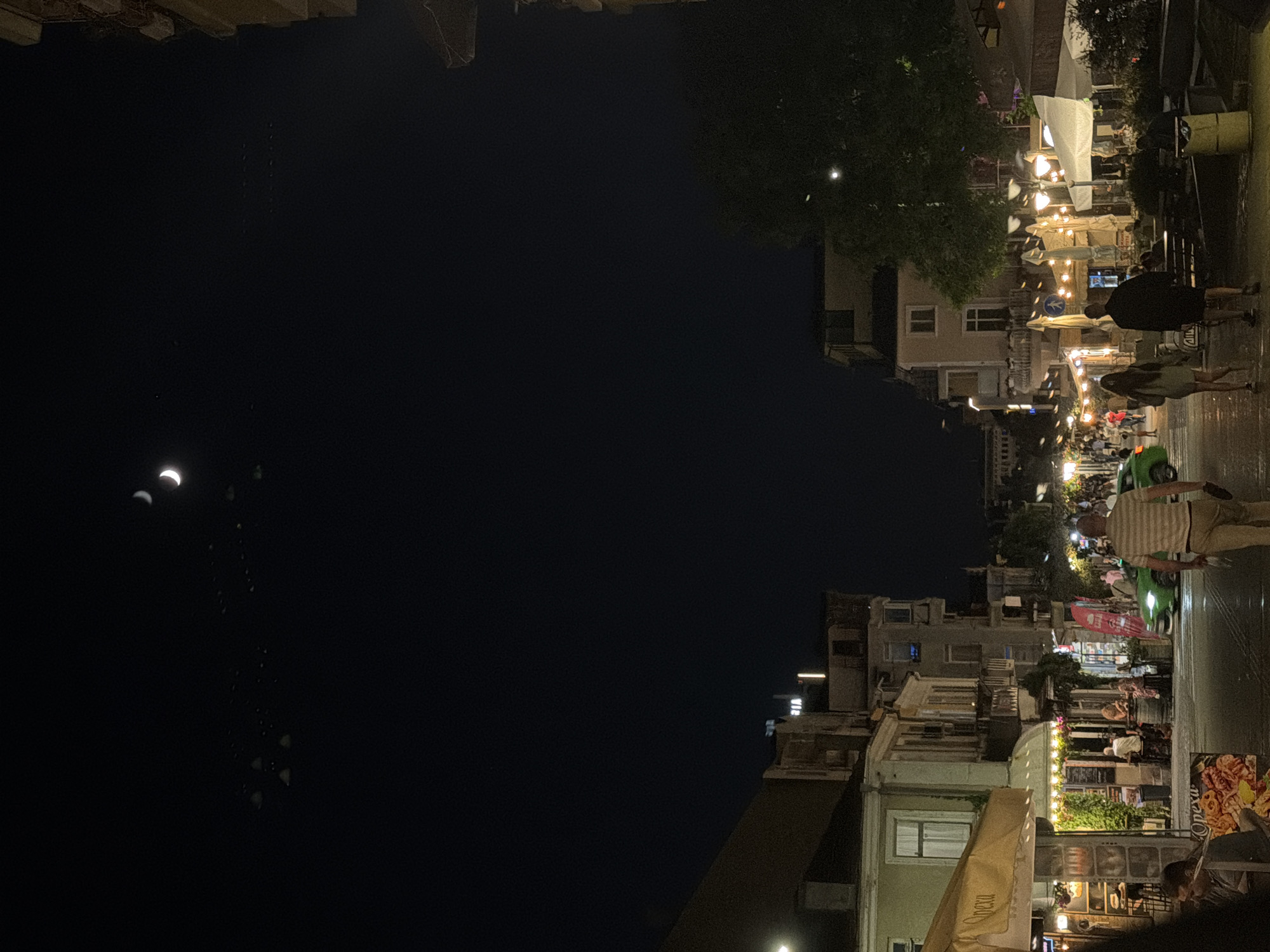 The image depicts a street scene at night in the city of Burgas, Bulgaria. The sky is dark with a crescent moon visible near the top center. A flock of birds is in flight across the sky. The street is lined with buildings, many of which appear to house businesses like restaurants and cafes, indicated by illuminated signage and outdoor seating areas. People are walking along the street, and some are seated at tables. A green car is driving down the street. The overall atmosphere is lively and urban, with artificial lights illuminating the scene.