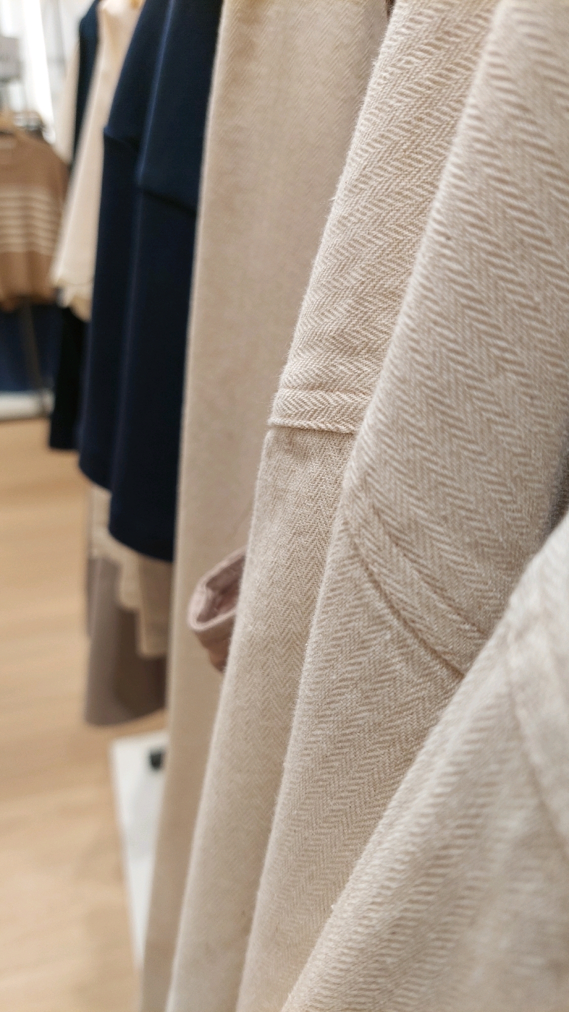 A close-up, slightly angled shot showcases several garments hanging on a rack, likely in a retail setting. The focus is on the textured fabrics, particularly a beige material with a herringbone weave that dominates the foreground. A dark blue garment hangs behind the beige one, and another beige garment is visible further back. The lighting is soft, highlighting the textures and folds of the clothing. The background is blurred, suggesting a shop interior with wooden flooring. The image does not depict any specific location within Bekasi, Indonesia.