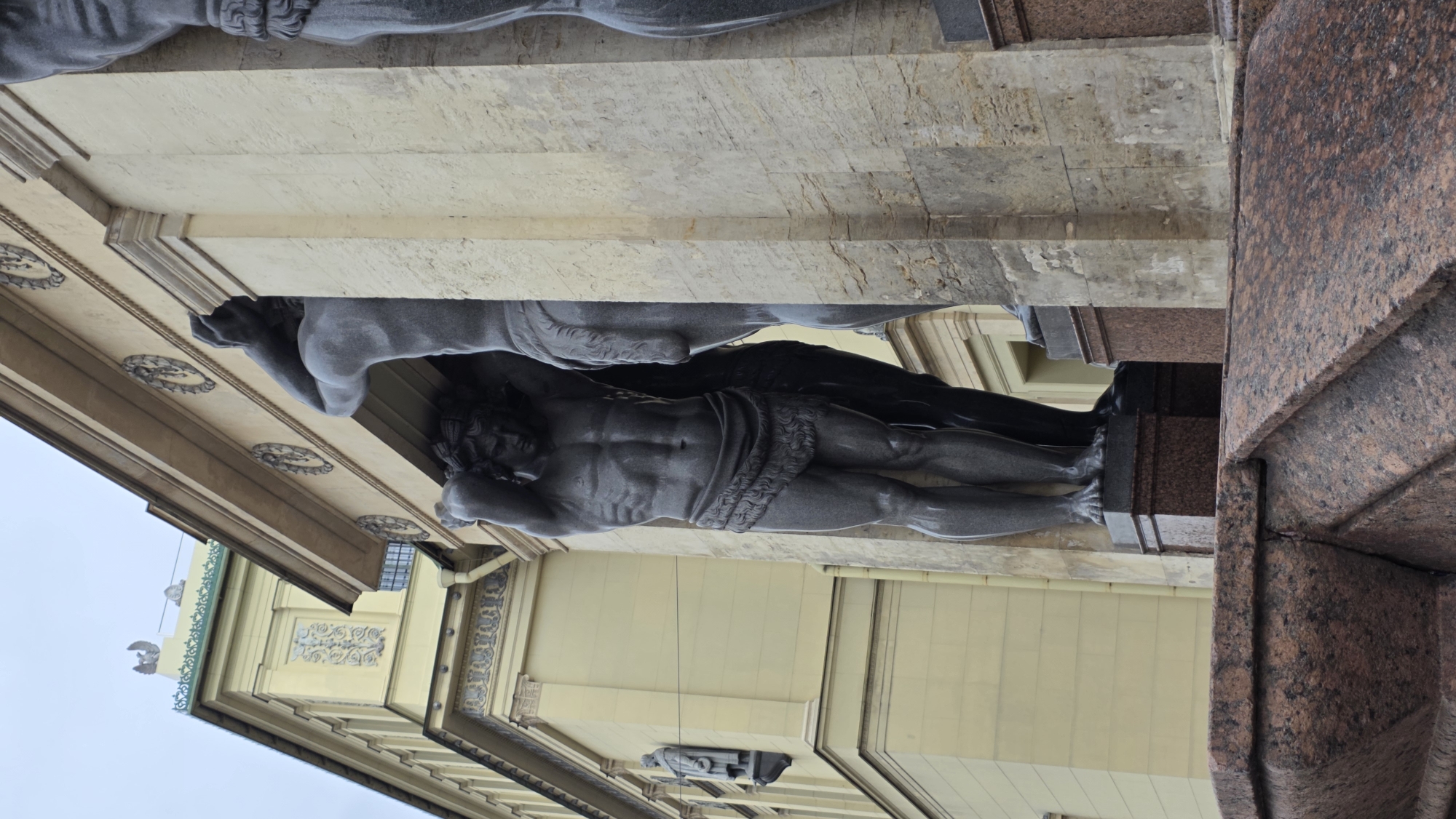 The image shows one of the Atlantes of the Winter Palace in Saint Petersburg, Russia. The gray granite statue of a muscular man is supporting a portico, with the building's facade in the background. The statue is part of a larger group that decorates the Hermitage Museum.