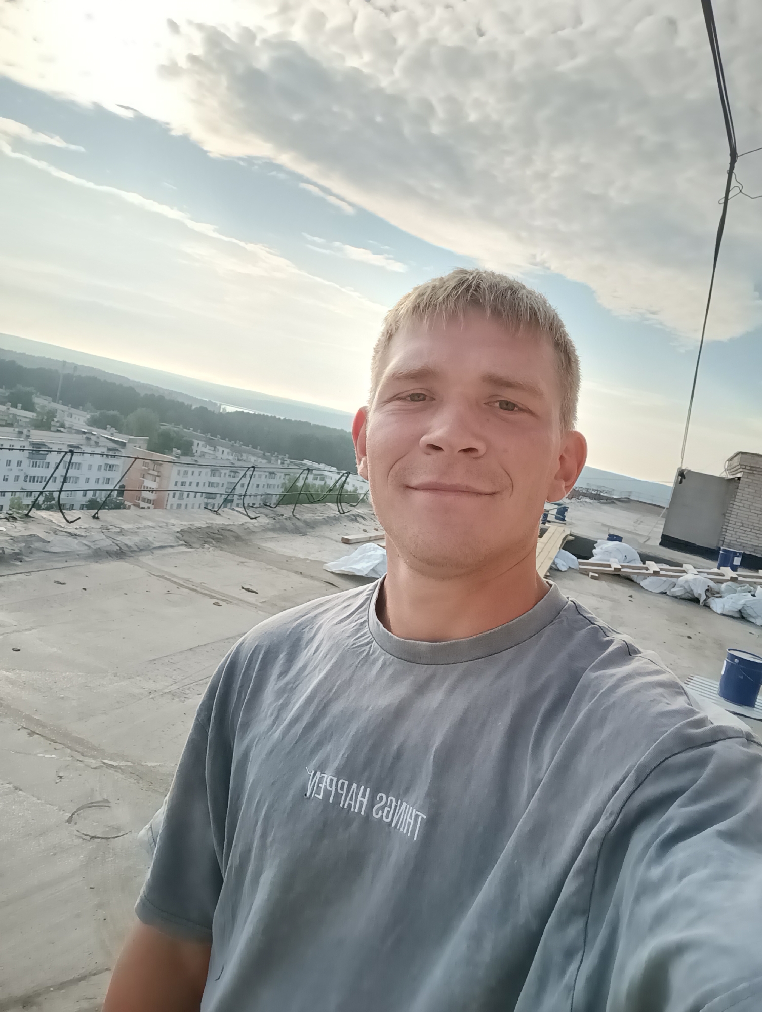 A man with blond hair and a gray t-shirt is taking a selfie on a rooftop. He is smiling at the camera. In the background, there is a cityscape with buildings and trees, under a cloudy sky. There are also some construction materials on the rooftop, including bags of materials, wooden planks, and blue buckets. The man's t-shirt has some text on it that appears to be in reverse.