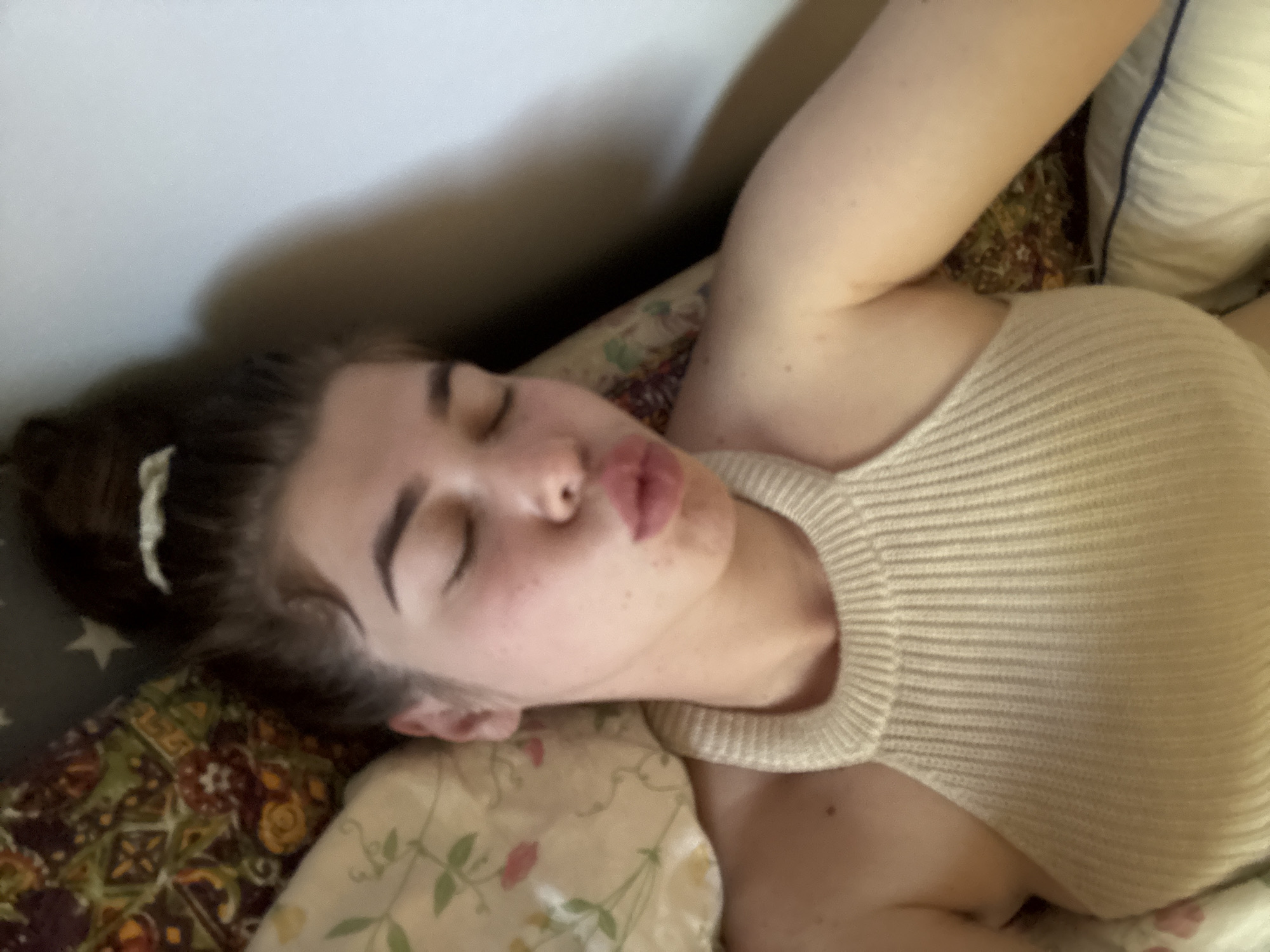 A young woman with her eyes closed and her lips puckered lies on a bed. She is wearing a cream-colored knit halter top. Her hair is pulled up into a bun. The background shows patterned pillows and a plain wall. The image appears to have been taken indoors.