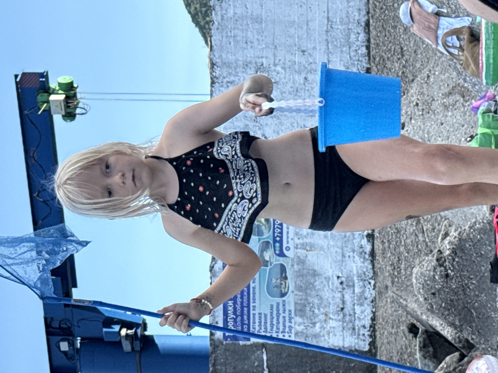 A young girl with blonde hair, wearing a black and white patterned bikini top and black bottoms, is pictured outdoors. She is holding a blue bucket in her right hand and a blue fishing net on a pole in her left hand. The girl appears to be looking towards the camera, with a neutral expression. The setting appears to be a seaside location, with a concrete wall and a body of water visible in the background. There are also some structures that suggest a pier or a dock, including a green crane. The sky is a clear blue, indicating a sunny day. The image is taken from a slightly low angle, and the girl is positioned in the center of the frame.
There is text visible on a poster in the background, which appears to be in Russian. The text mentions various activities such as "boat trips", "banana boat rides", "fishing", "diving", "jet skis", "catamarans", "water skiing", and "SUP boards". It also includes a phone number and a website address. The overall impression is that of a vacation or a day spent at a recreational beach area.
The orientation of the image is rotated, suggesting it may have been taken with a mobile phone camera. The lighting suggests it is daytime, likely midday due to the bright sunlight.