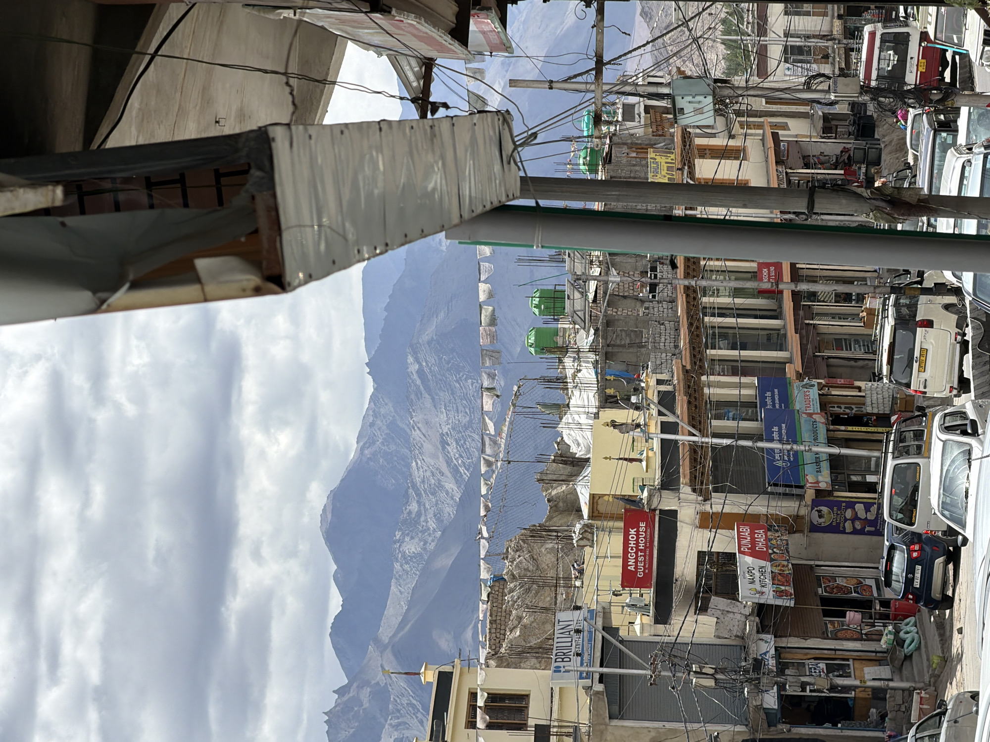 This is a ground-level, medium shot of a busy street scene in Leh, India, with majestic, snow-capped mountains in the background under a cloudy sky. The foreground is dominated by the side of a building with corrugated metal sheeting and exposed wiring, partially obscuring the view. Below this, a street is lined with shops and guesthouses. Signs visible include "BRILLIANT SANITARYWARE," "ANGCHOK GUEST HOUSE," "NAKPO PUNJABI KITCHEN," and "TRADER'S." Several vehicles, including cars and a red and white minibus, are parked on the street. Prayer flags are strung across some of the buildings. The architecture appears to be a mix of traditional and modern styles. Electrical wires are densely strung throughout the scene, a common feature in many urban areas. No people are clearly identifiable or engaged in any specific actions. The overall impression is a bustling commercial street in a high-altitude, mountainous region.