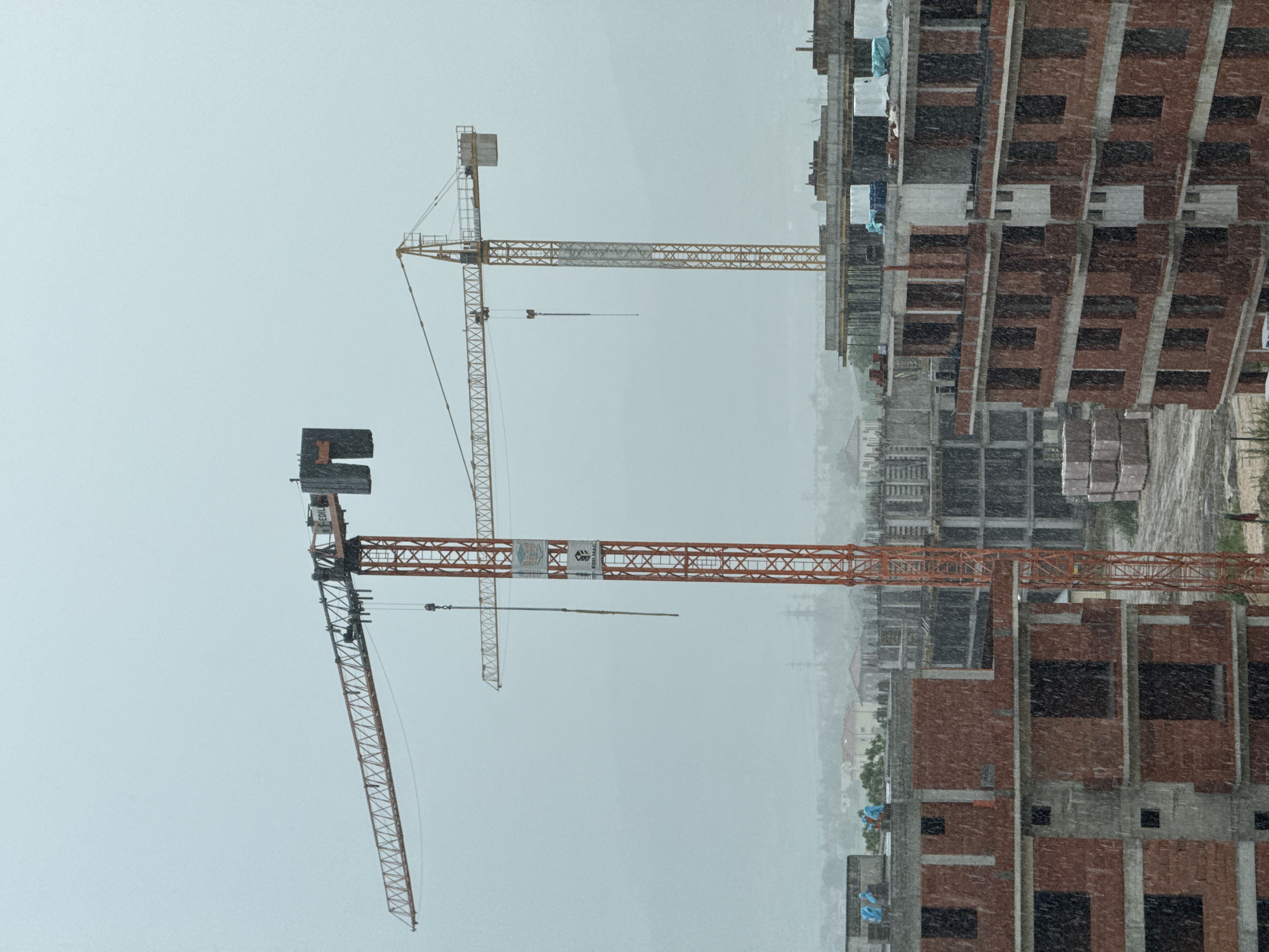 The image depicts a construction site in Plovdiv, Bulgaria, during a snowstorm. Two cranes are visible against a gray sky, standing adjacent to the unfinished brick structures of buildings under construction. The scene is shrouded in snowfall, reducing visibility. There are no people visible in the scene.