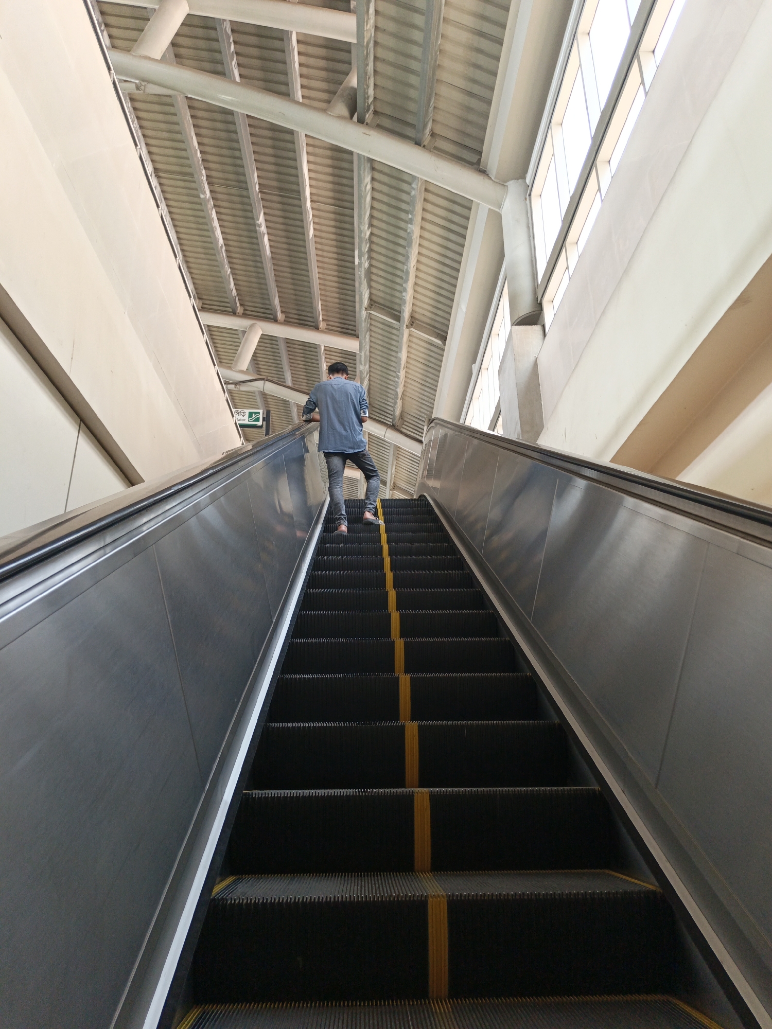 A person in a blue shirt and jeans is riding an escalator upwards. The escalator is black with yellow lines. The surrounding architecture includes a beige wall, a metal ceiling structure, and windows. A green exit sign is visible. This is likely taken in a public transportation hub such as a metro or a train station in Dhaka, Bangladesh.