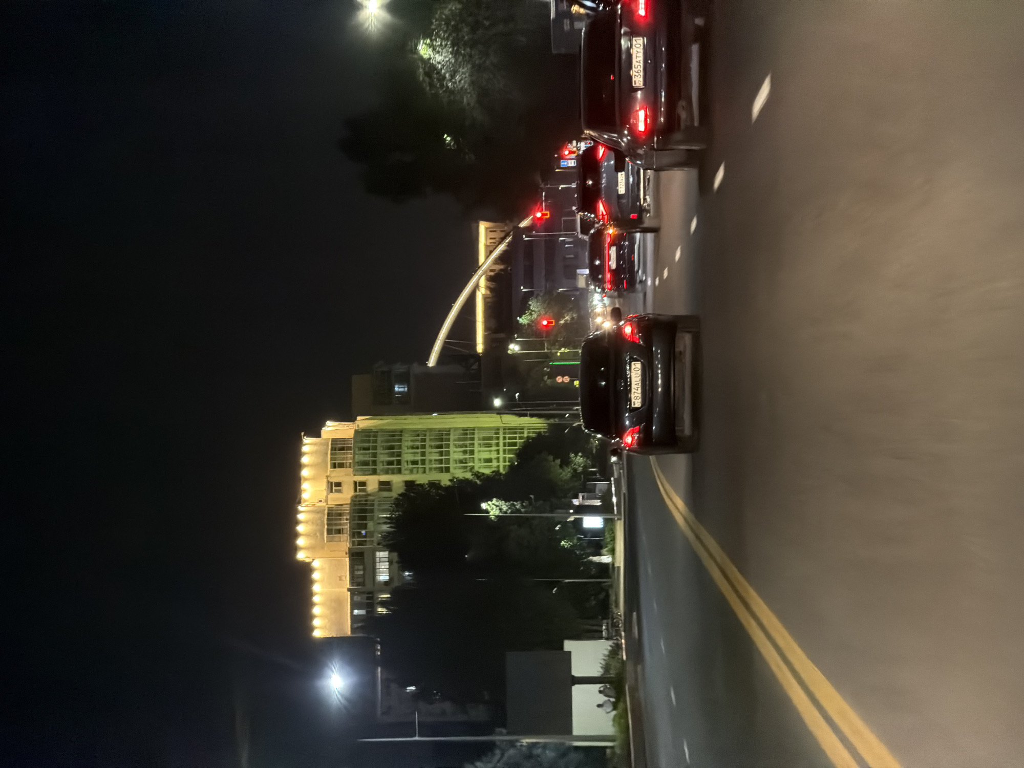 The image depicts a nighttime scene of a city street in Astana, Kazakhstan, specifically along Dostyk Avenue. The perspective is from within a moving vehicle, looking forward. Several cars are visible on the road, traveling in the same direction. The road is paved and has double yellow lines marking the lanes. Traffic lights are illuminated red in the distance, indicating a stop.
Dominating the background on the left is a modern, multi-story building illuminated with warm yellow lights, its facade featuring a unique, stepped design along the roofline and green-tinted glass on the lower floors. Adjacent to this building, trees are silhouetted against the night sky and the building's light. Further in the distance, an architectural bridge with glowing arches spans across the view. Streetlights are visible along the sides of the road, casting pools of light.
On the right side of the image, more trees are visible, also dark against the night. The rear taillights of the cars in front glow red. One car in the foreground on the right displays a license plate with Cyrillic characters and the number "365ATY 01". Another car ahead of it shows a license plate "723KGN". A car in the lane to the left has the plate "774ALU 01".
On the left side of the street, a small group of people are seated on a bench near a tall, illuminated billboard or sign structure. The overall atmosphere is that of a typical urban night, with vehicular traffic and illuminated buildings.