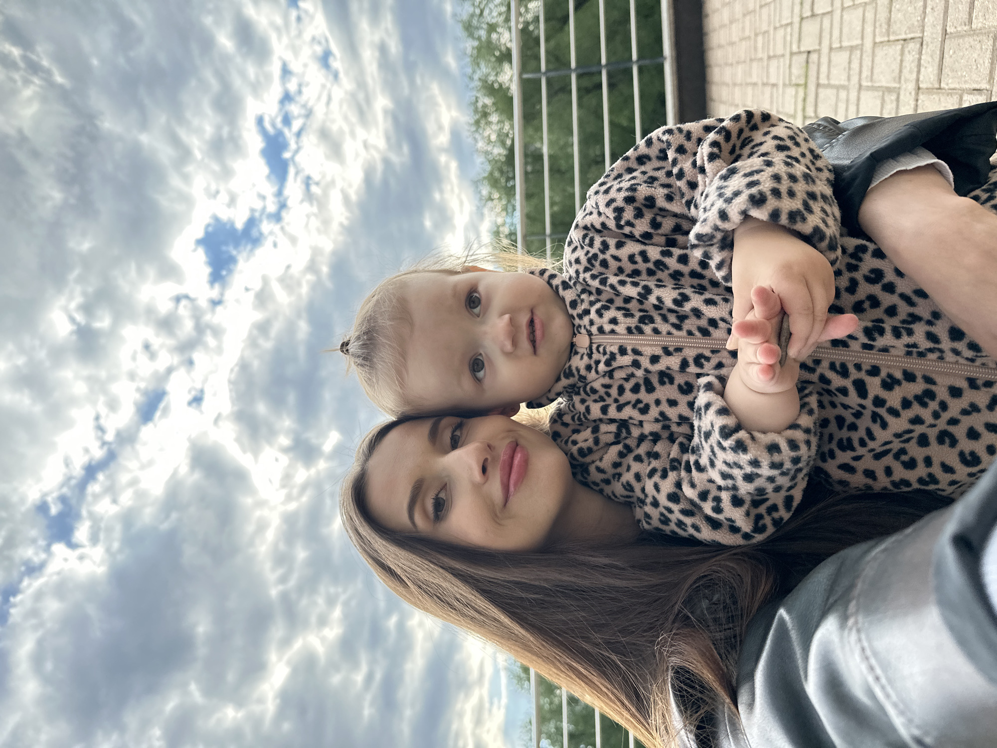 This image is a close-up, vertically oriented portrait, rotated 90 degrees clockwise, featuring an adult woman and a young child outdoors in Yekaterinburg, Russia. Both subjects are looking towards the camera, with the woman's head positioned slightly lower and to the left of the child's.
The woman has long, straight brown hair, with subtle makeup highlighting her eyes and lips, and a gentle, soft smile. The child, likely a toddler, has light blonde hair tied back in a small ponytail and large, curious eyes. The child is wearing a beige and black leopard-print zip-up fleece jacket. One of the child's small hands is visible, clutching a thin, dark object, possibly a twig or piece of debris. The woman appears to be wearing a dark, possibly grey or black, garment, partially visible on her shoulder and arm.
The background is dominated by a bright, cloudy sky with patches of clear blue visible between large, fluffy white and grey clouds. Below the cloud line, hints of a metal railing and green foliage from trees suggest an outdoor setting, potentially a park, a balcony, or an elevated walkway. A small section of light-colored paved ground or brickwork is also visible in the upper right portion of the image (due to rotation). The natural light indicates it is daytime, possibly a bright, overcast day or late afternoon. There is no visible text in the image.