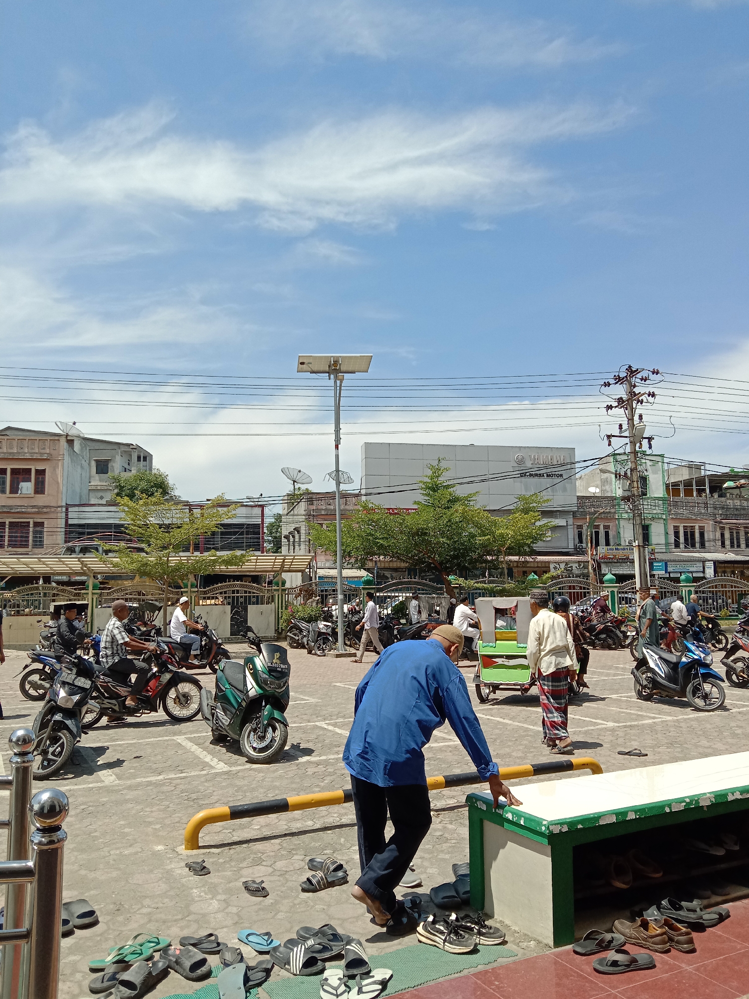 The image captures a street scene in Lhokseumawe, Indonesia, likely near a mosque or a public gathering area. The sky is blue with scattered white clouds. In the foreground, a man in a blue shirt and dark pants is bending over, his hand touching a green and white bench which serves as a shoe rack. Scattered on the ground around the bench are numerous pairs of sandals and shoes. To the left, a metal railing is visible.
The middle ground is a paved area with marked parking spots, where many motorcycles are parked and some are being ridden. Several individuals are on or near the motorcycles. One man is riding a green motorcycle, while another, in a blue shirt and a cap, is on a blue motorcycle. A person in a white shirt and a checkered sarong is walking away from a small, green and white cart. In the background, buildings line the street, including a structure with a prominent "YAMAHA" logo, indicating a dealership or service center. There are also satellite dishes visible on some of the buildings, and power lines crisscross the sky. Trees with green foliage are interspersed among the buildings, adding to the urban landscape. The overall impression is a busy, sunny day in a commercial or community hub.