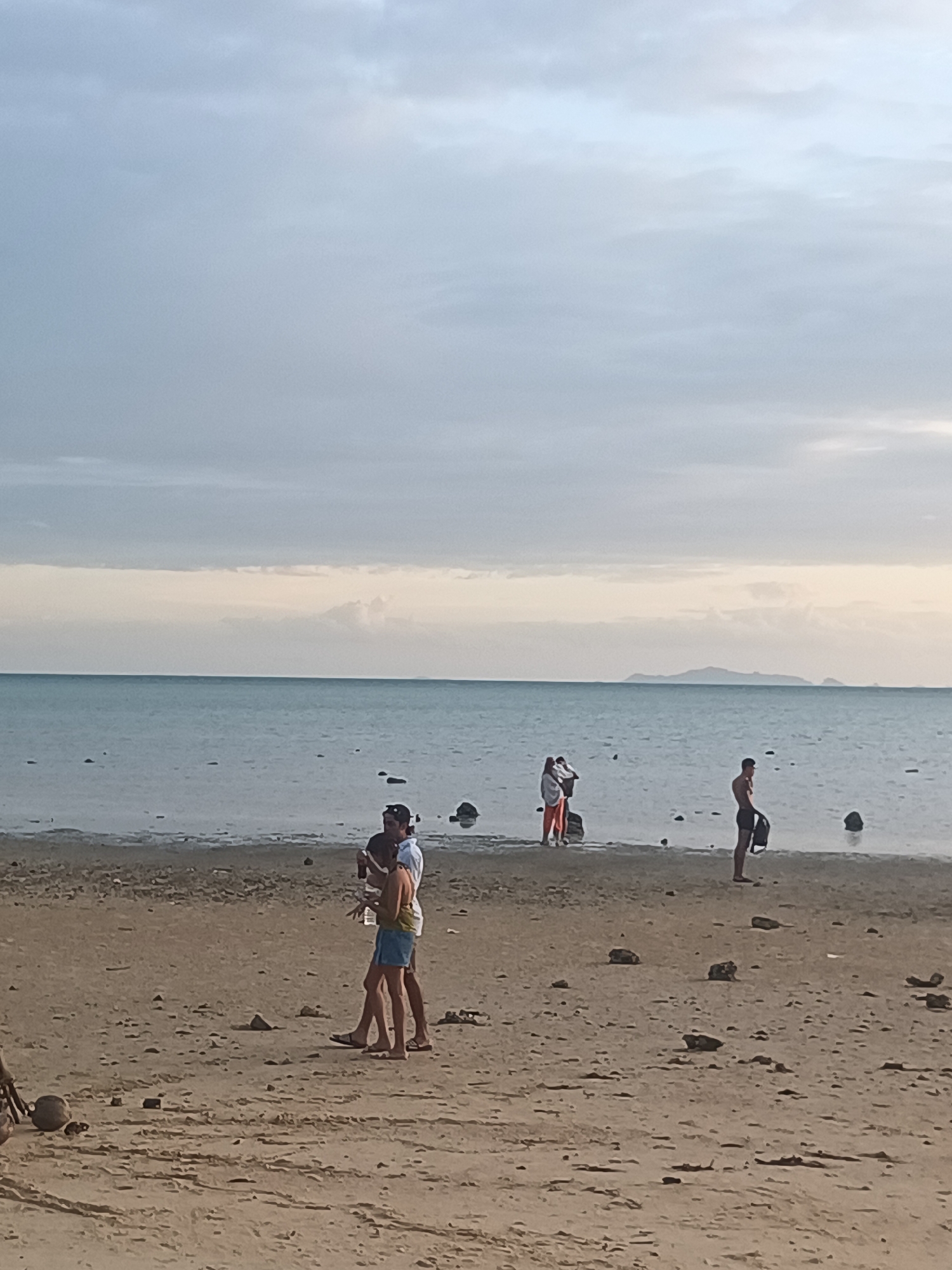 The image depicts a beach scene in Tambon Ko Phangan, Thailand. In the foreground, two individuals, a male and a female, are walking on the sandy shore. The male is wearing a white shirt and blue shorts, while the female is wearing an orange hoodie and shorts. They appear to be engaged in conversation. Further in the background, another male is standing by the water's edge, holding a bag. In the distance, a group of people are visible near the water, with one individual appearing to be holding a camera. The sea stretches out to the horizon under a cloudy sky. The beach is littered with small stones and debris.