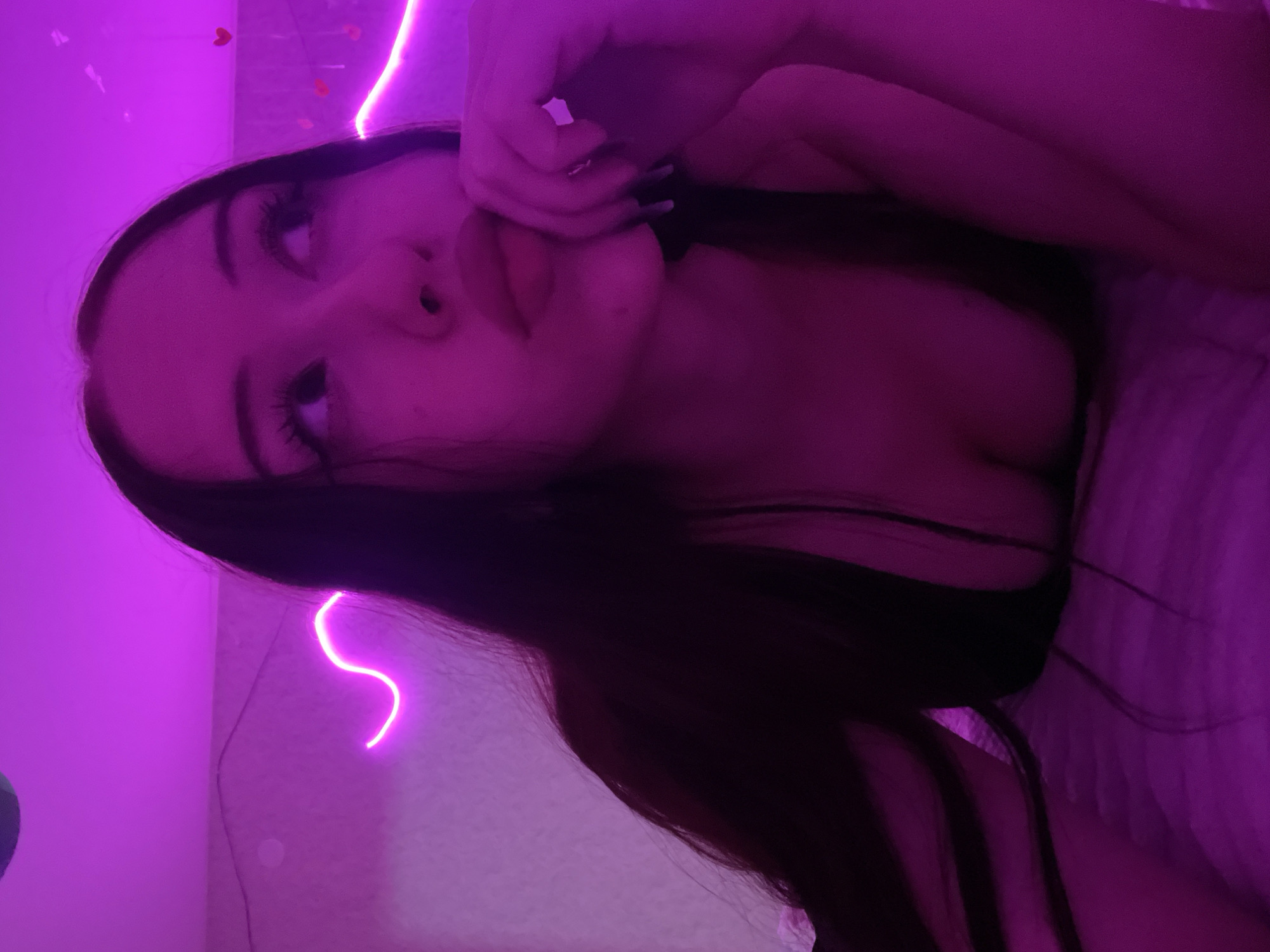 The image shows a young woman with long, dark hair lying on a bed. She is looking upwards and to the right, with her hand touching her chin. The room is lit with pink neon lights, one of which is a wavy line on the wall behind her, and another is a string of small hearts. The woman is wearing a dark top that reveals her cleavage. The bedding is textured and appears to be a light color. The image is taken from a slightly low angle. There is no information in the image to suggest it is located in Votkinsk, Russia.