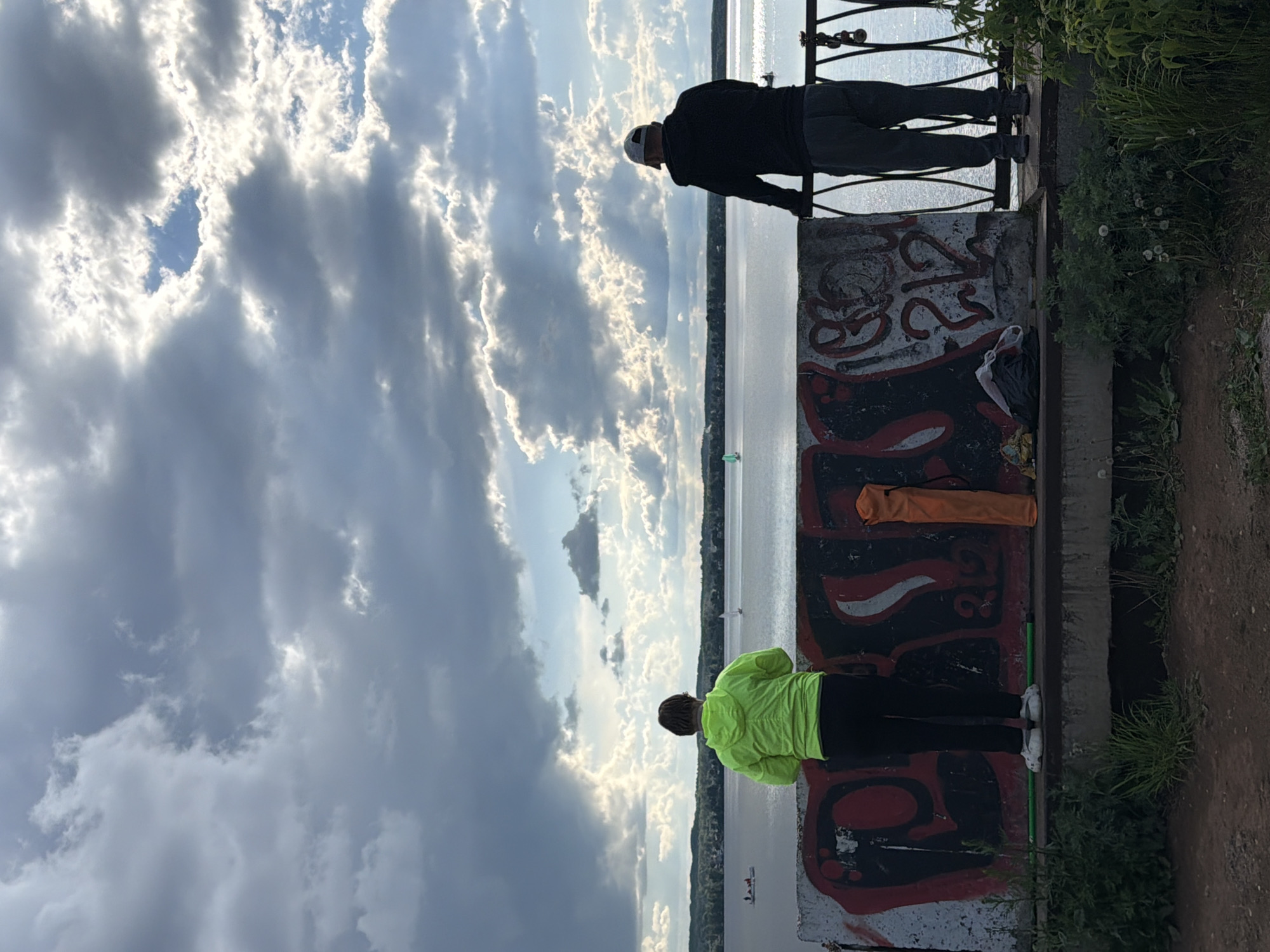 The image depicts a waterfront scene in Votkinsk, Russia. Two individuals are standing on a concrete platform overlooking a body of water, possibly the Votkinsk Reservoir. The platform features graffiti and is bordered by a metal railing. One person is wearing a black jacket and light-colored pants, leaning on the railing. The other person, clad in a neon yellow jacket and dark pants, stands closer to the graffiti-covered wall. An orange roll-up bag and a dark bag rest against the wall near the individuals. Vegetation grows along the edge of the platform. The sky is filled with dramatic clouds, suggesting either a stormy or sunset-lit atmosphere. In the water, several small boats are visible.