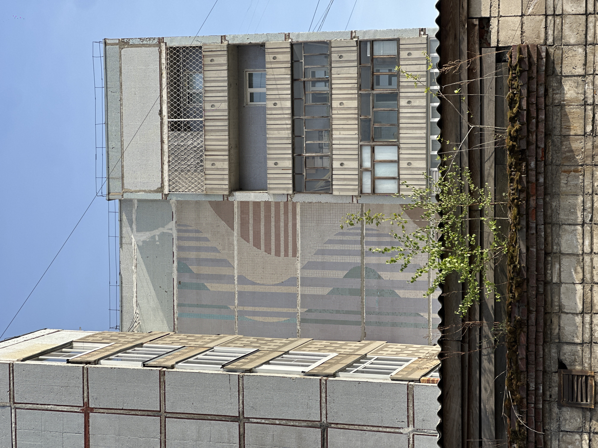 An eye-level, rotated image depicts an exterior view of two adjacent buildings. The buildings are both constructed of concrete blocks, each unit having a tiled surface. The building on the left has rows of rectangular windows covered with a brown, wooden-like material. The top floor features a large mosaic design of brown, white, and blue hues. The windows of the upper level are smaller and more numerous than the bottom level.
The building on the right, with a vertical orientation, shows a rougher construction than its left counterpart. Its wall shows a vertical corrugated material and an exposed brick portion. Vines and moss are growing on the side of the wall.
The scene is set against a light blue sky.