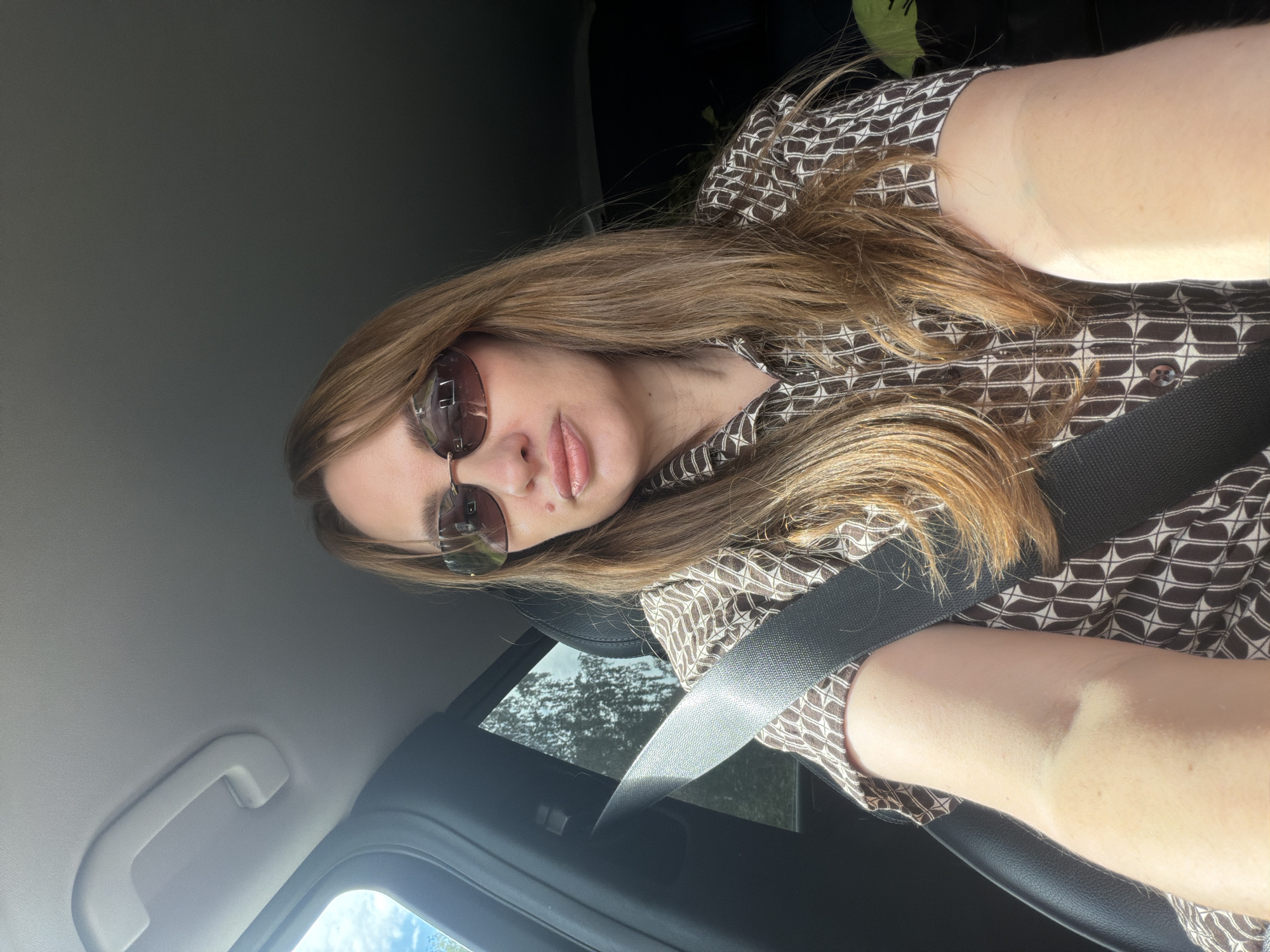 A woman with long, brown hair and sunglasses is sitting in the driver's seat of a car, looking at the camera. She is wearing a brown and white patterned shirt and a seatbelt. The car interior is visible, including the dashboard and a side window showing trees and sky. The scene is well-lit, suggesting it is daytime. The location is Poletaevo 1th, Russia.