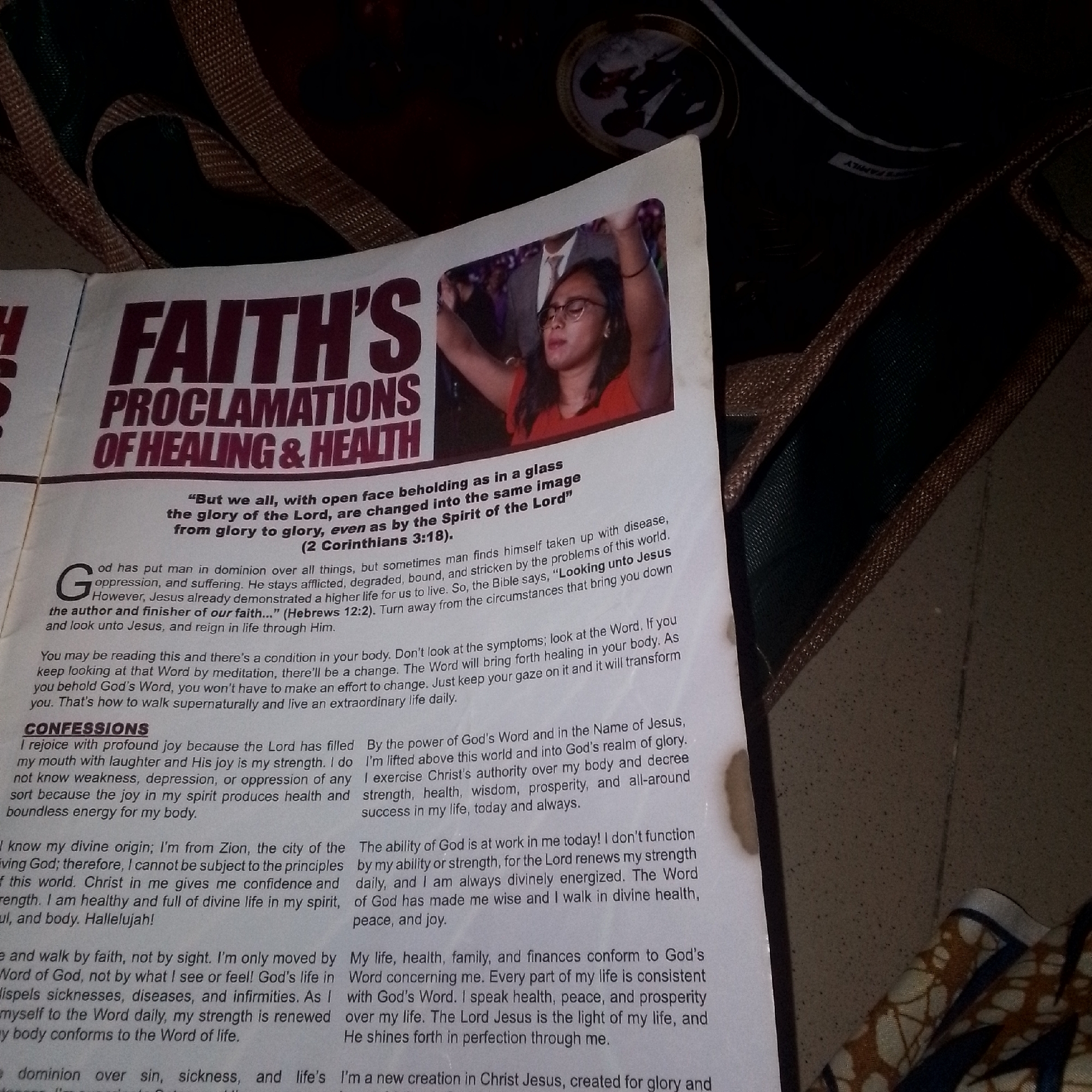 The image shows a close-up view of a printed document with large, bold text and an accompanying photograph. The title of the document appears to be "FAITH'S PROCLAMATIONS OF HEALING & HEALTH." The photograph is of a young woman wearing glasses and an orange top, with her hands raised in the air and her eyes closed. She is looking upwards. The document contains text in a smaller font below the title and the photograph. The scene is lit, and the background consists of a dark, textured surface, possibly a bag or upholstery, and a lighter, speckled surface, likely a table or floor. The document is positioned on top of these surfaces. The image does not contain any visual information that could indicate its location in Jalingo, Nigeria.