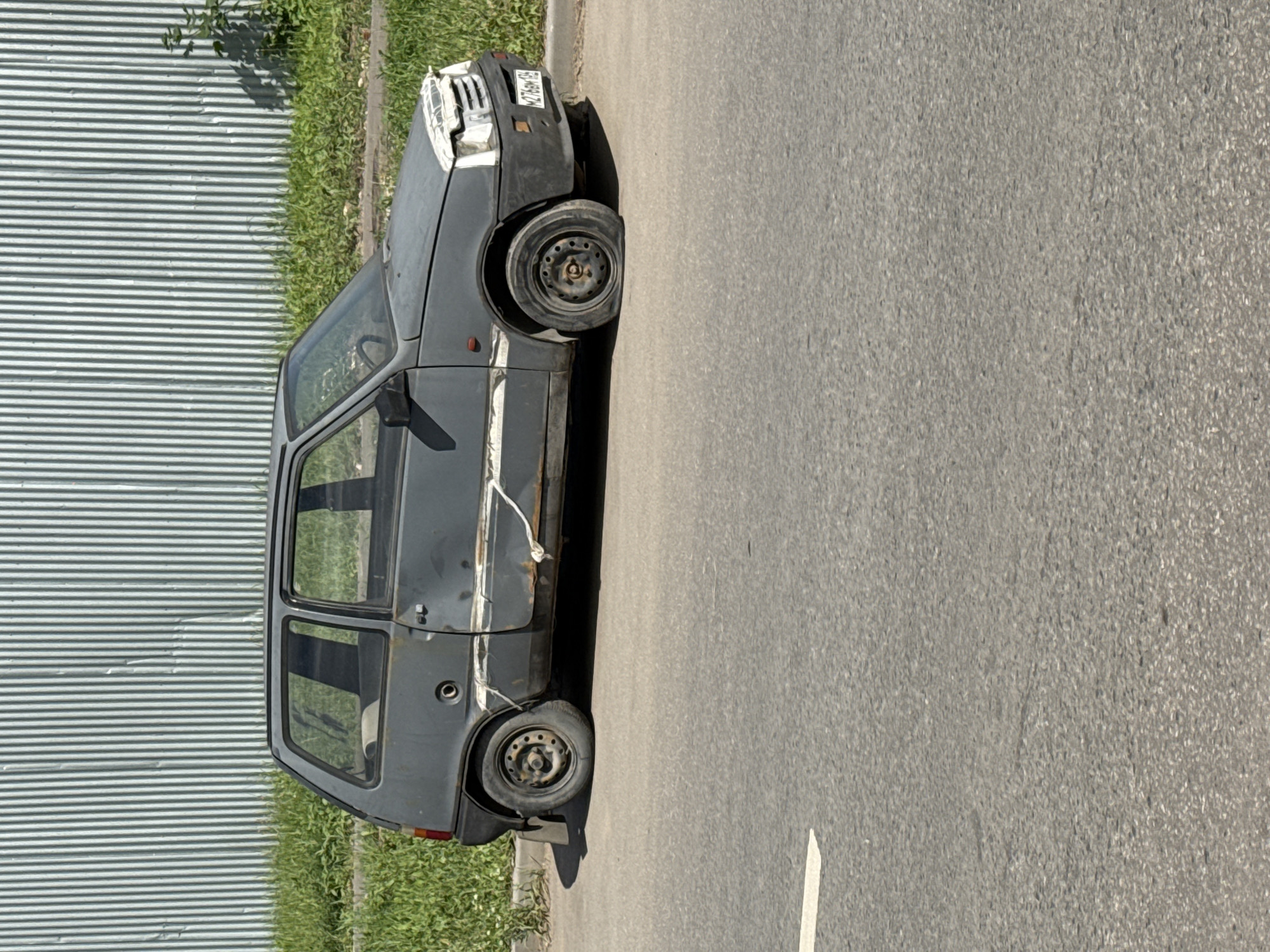 The image shows a dark gray, old, heavily damaged car parked on the side of a paved road. The car is oriented vertically in the frame, with its front end pointing upwards. The left side of the car faces a corrugated metal fence and a strip of green grass. The right side of the car faces the road, which appears to be asphalt with a white line marking.
The car's body shows significant wear and tear, including rust along the side panels and a dented fender. One of the front tires appears to be flat or missing. The windshield is dirty, and the side windows are tinted. The license plate is visible on the front of the car, but the text is blurry.
The scene suggests a neglected or abandoned vehicle. The time of day appears to be daytime, likely sunny, as indicated by the shadows cast by the car. The setting is urban or suburban, given the presence of the road and fence. No people are visible in the image.
The overall impression is one of disuse and decay. The location context provided is Moscow, Russia.