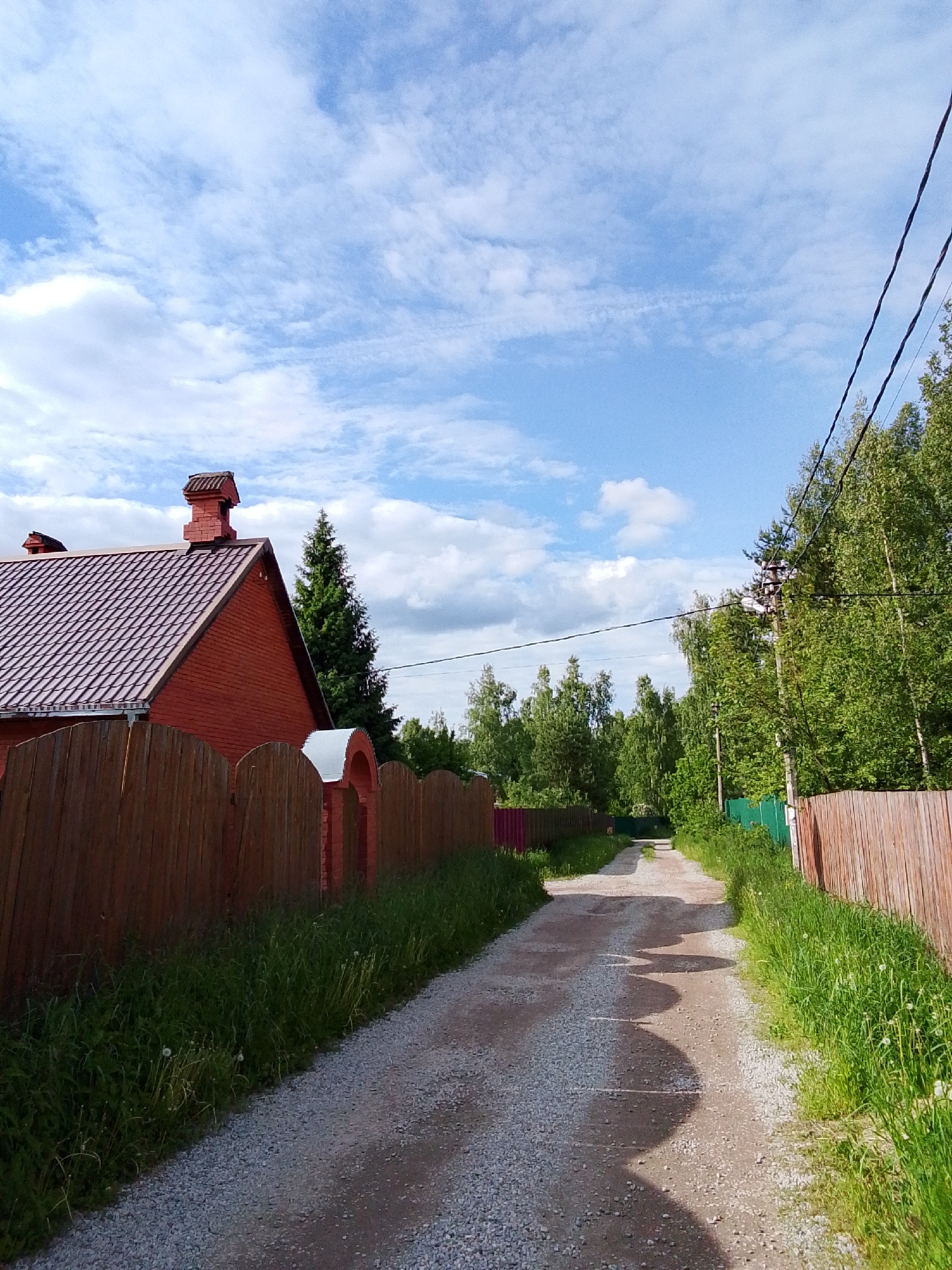 The image shows a gravel road in Снопок Новый, Russia. The road is bordered on both sides by wooden fences, with a red brick house and an arched gate visible behind the fence on the left. Tall trees and green vegetation line the road, and a blue sky with scattered clouds is overhead. Overhead power lines are present on the right side of the image. The road has several patches of darker gravel.