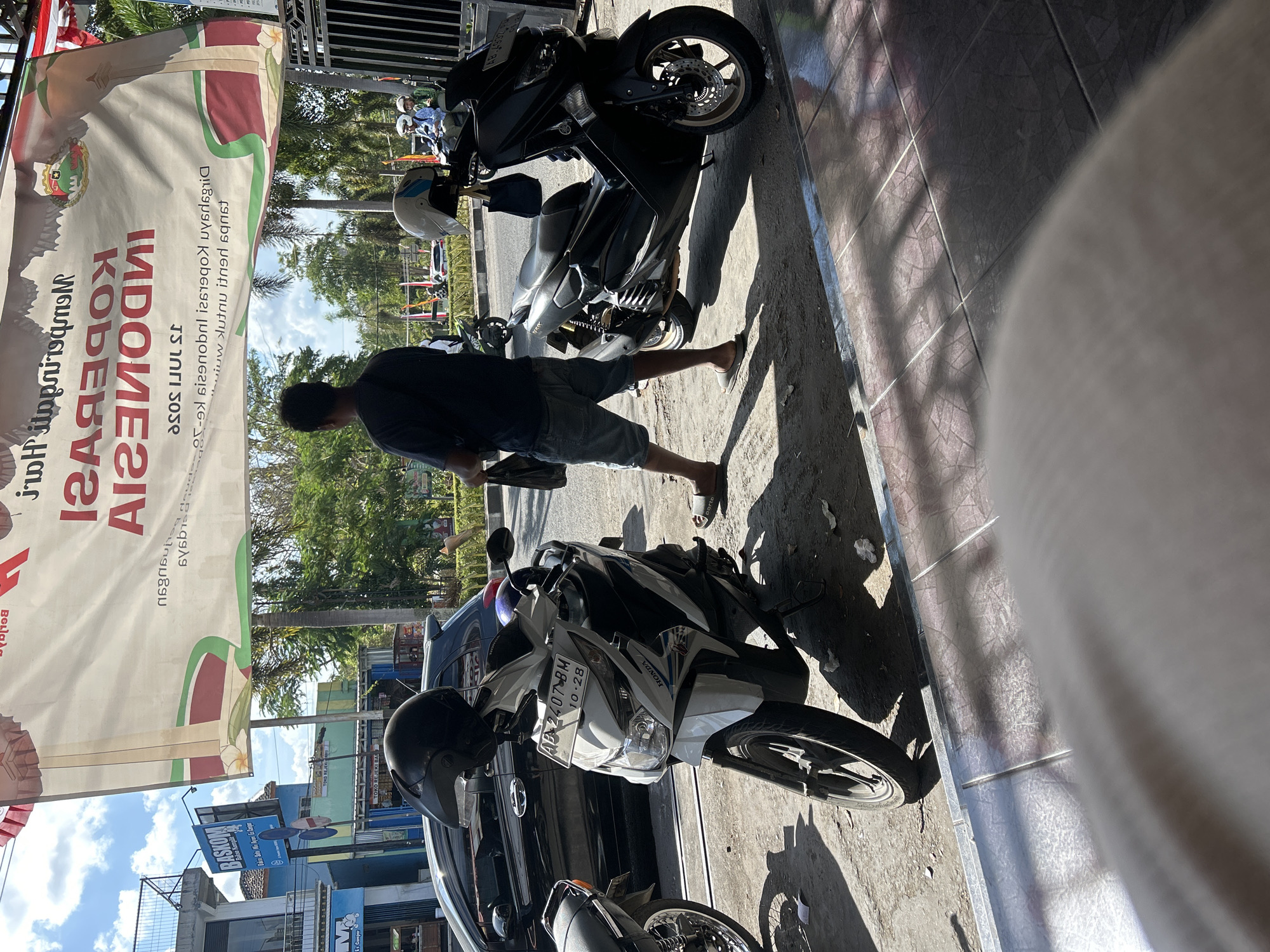The image shows a street scene in Wonosari, Indonesia, on a sunny day. In the foreground, a white Honda scooter is parked, facing left. A black helmet is placed on its handlebars. To the right of the Honda scooter, a black Yamaha NMAX scooter is parked, also facing left. A blue bag hangs from the handlebars of the Yamaha.
A man wearing a dark t-shirt and denim shorts stands between the two scooters, facing away from the camera. He is holding a black plastic bag in his right hand. His left hand is not visible. He is wearing flip-flops. Behind the scooters, a black car is partially visible.
In the background, a large banner with text and an image is displayed. The text on the banner appears to be in Indonesian, with some of it reversed. There are trees and buildings visible behind the banner. A sign for "BASKOM" is visible on a blue building to the left. Further in the background, there are more buildings and trees. The sky is blue with some clouds. The ground in the foreground is paved with tiles and asphalt.