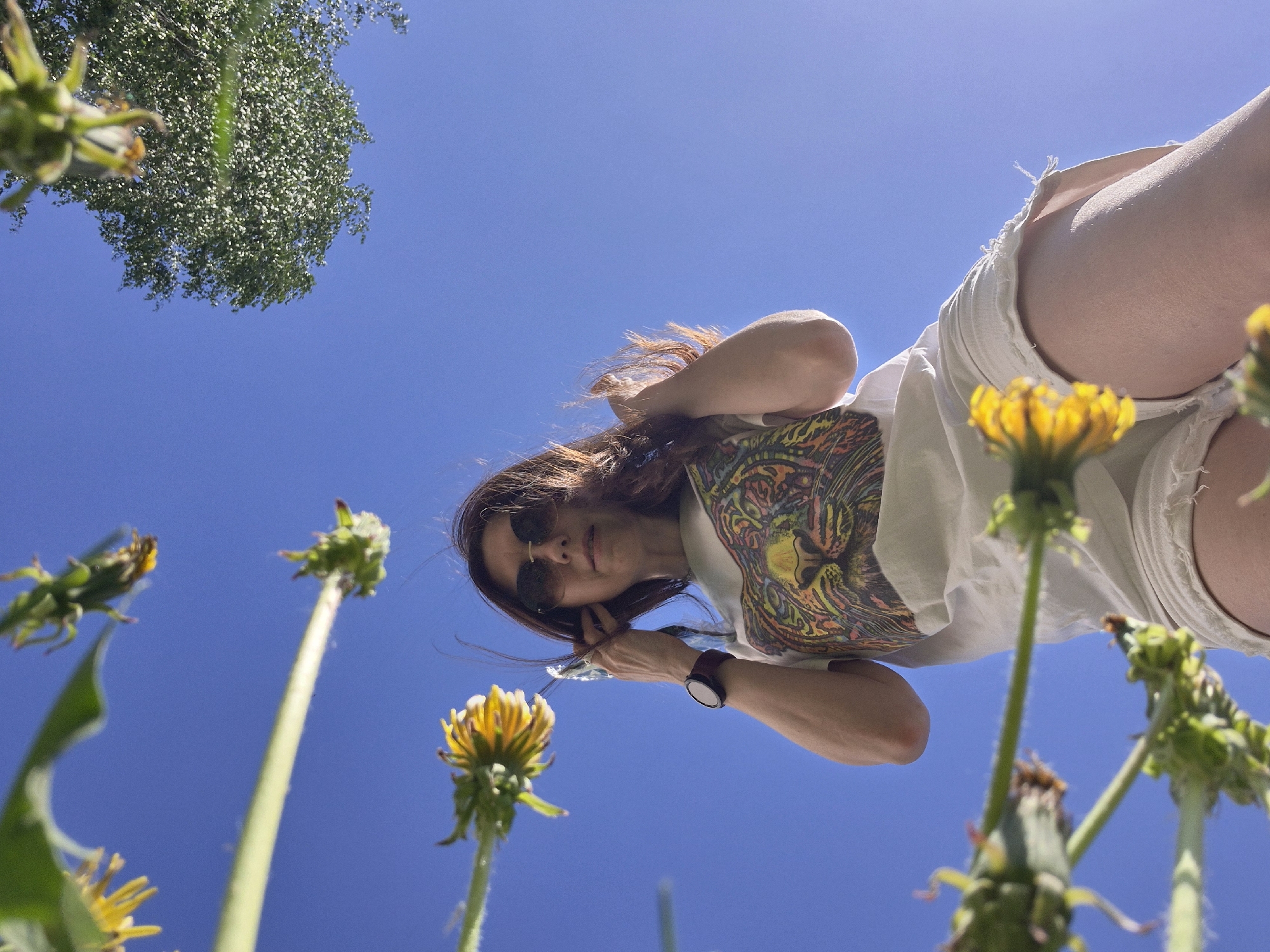 A low-angle shot looking up at a woman standing outdoors. She is wearing sunglasses, a patterned shirt, and white shorts. Her hair is brown. She is holding her phone and wearing a watch on her left wrist. The background is a clear blue sky with some greenery, including dandelions, visible around her. There is also a tree branch. The location is undetermined.